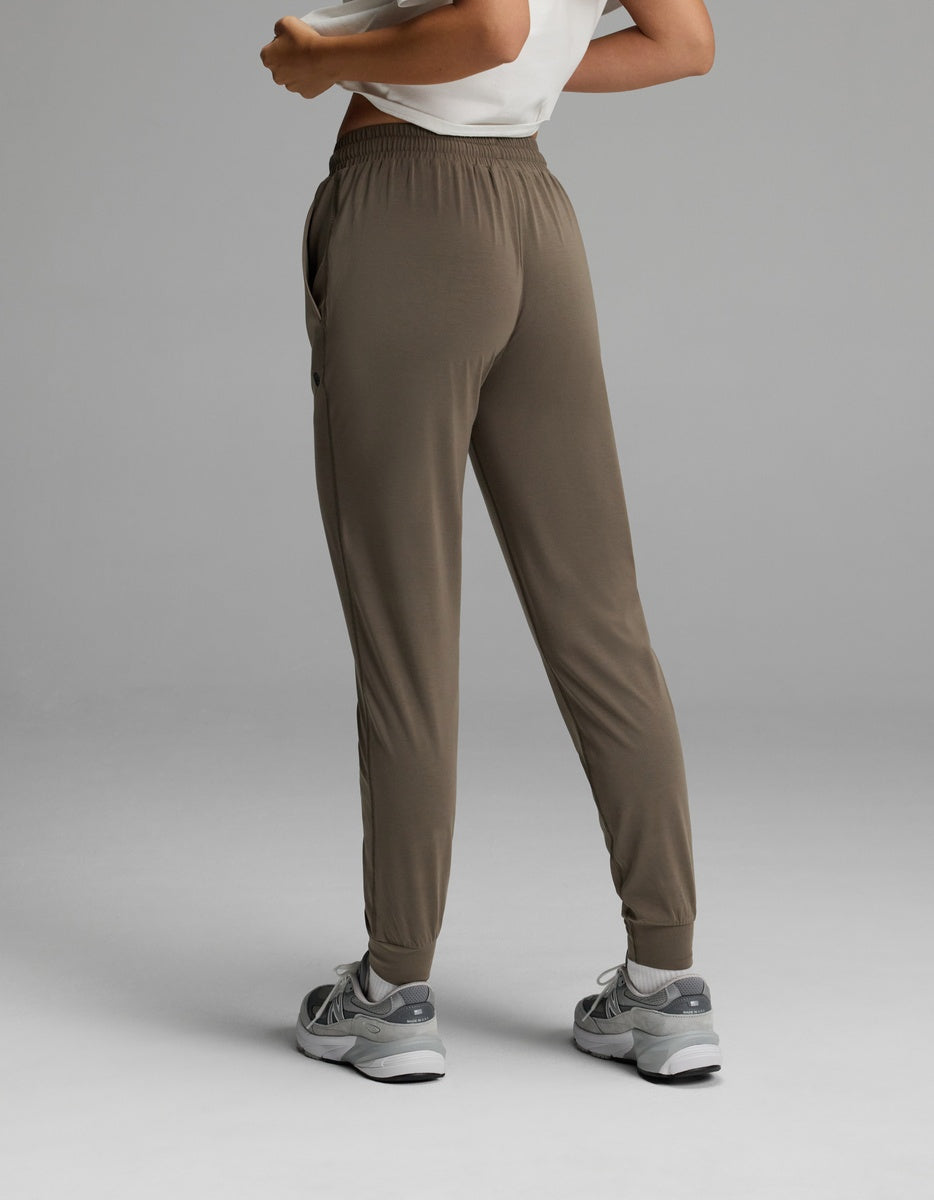 A person is seen from behind wearing the Cozy Earth Women’s Studio Jogger in taupe, paired with gray sneakers and a white top. The image highlights the fit and style of the pants against a plain gray background.