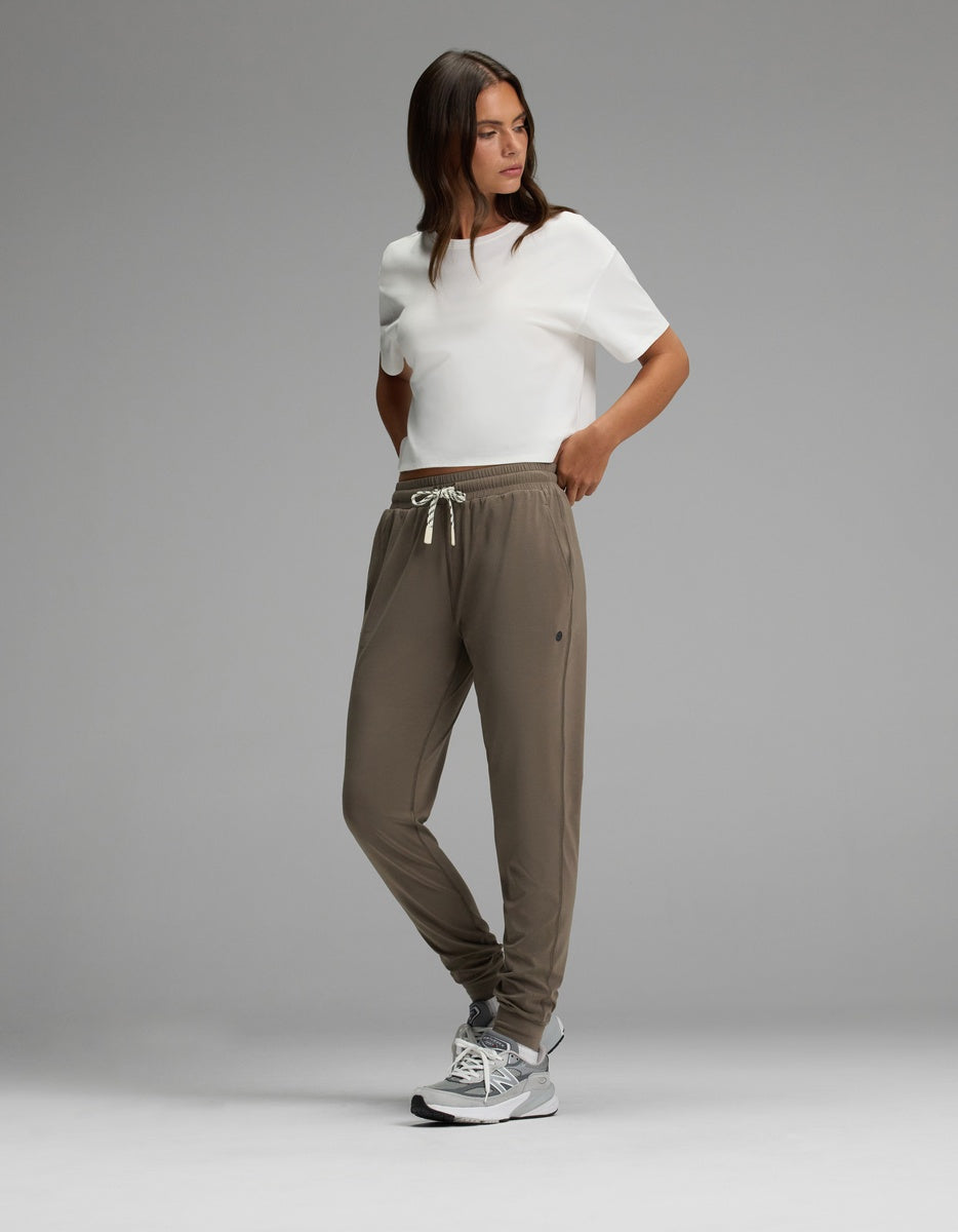A woman stands against a gray background wearing Cozy Earth’s Women’s Studio Jogger in taupe with a drawstring waist, paired with a white short-sleeve shirt and gray athletic shoes. Her hands are in her pockets and she is looking to the side.
