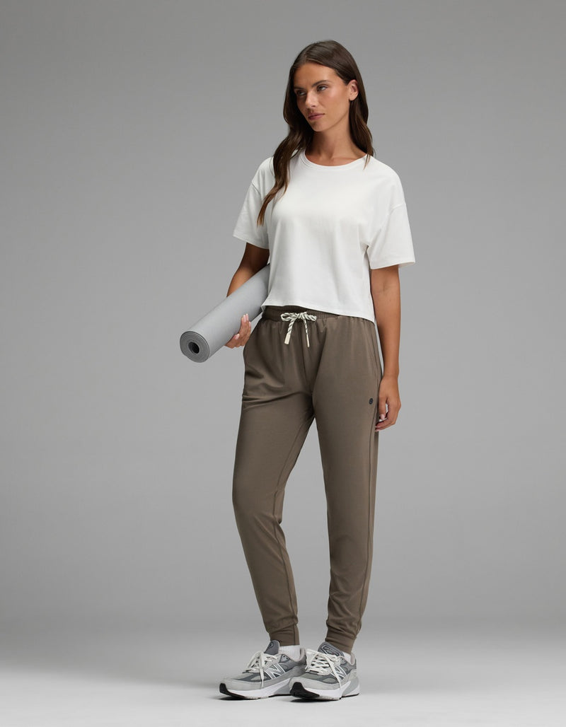 A woman with long brown hair stands on a gray background, wearing Cozy Earth’s Women’s Studio Jogger in taupe and a white T-shirt. She holds a rolled-up yoga mat and wears sneakers.