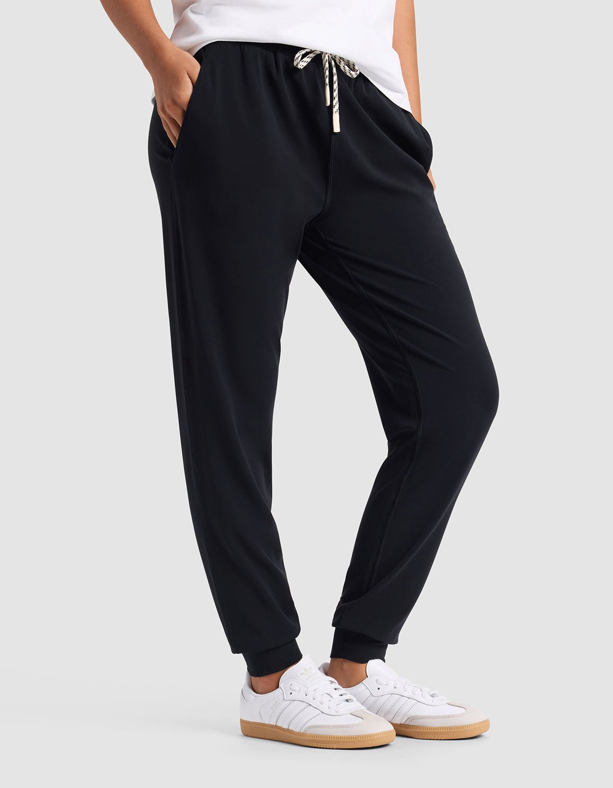 A woman wears Cozy Earth Women's Studio Jogger—black with a drawstring waist—paired with white sneakers and a white top, standing hands-in-pockets against a plain light gray background.|Color:Jet Black