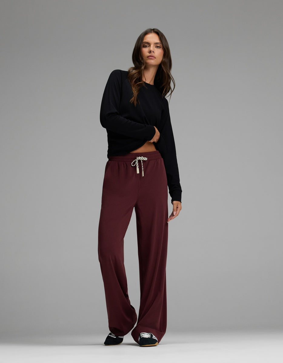 A woman with long brown hair and a relaxed expression stands against a gray backdrop, wearing Cozy Earth’s Women’s Studio Wide Leg Pant in burgundy, paired with a long-sleeve black top and black shoes.