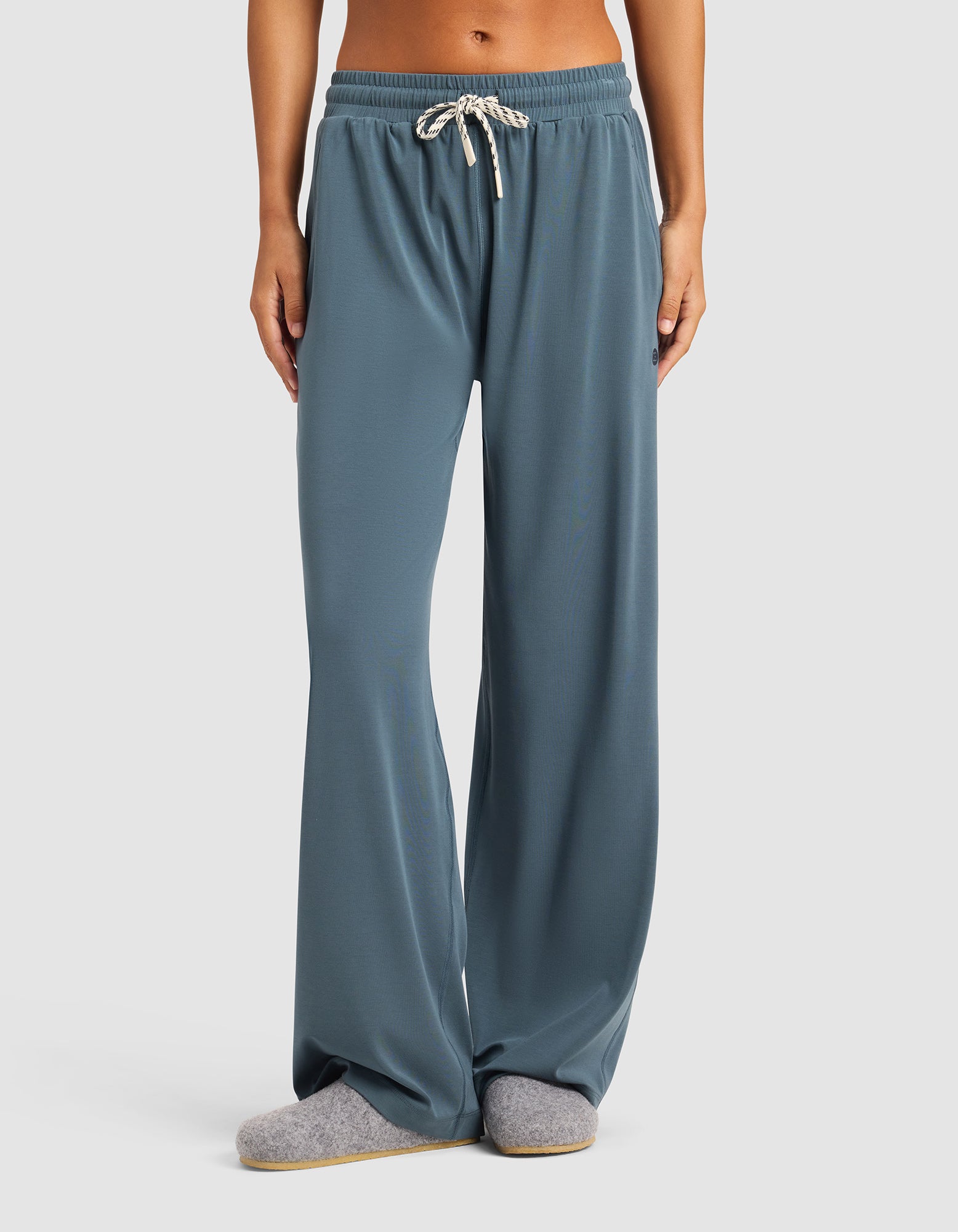 A person wears Cozy Earth's Women's Studio Wide Leg Pant in blue-gray, paired with gray slippers, standing against a plain light background. Only the lower half of the body is shown.