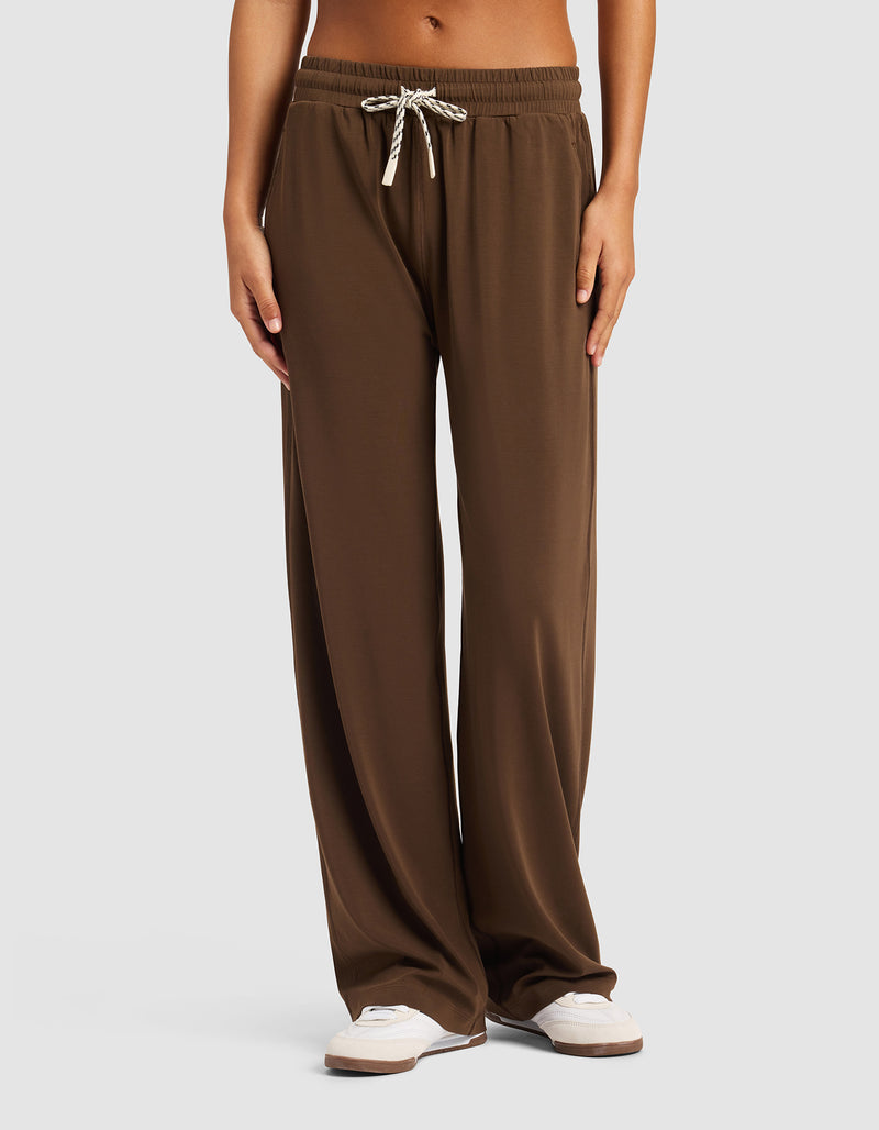 A person is standing against a light gray background wearing Cozy Earth's Women's Studio Wide Leg Pant in brown with white sneakers, hands relaxed by their sides. The pants feature a loose fit and drawstring waist.