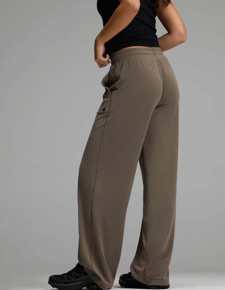 A woman models the Cozy Earth Women’s Studio Wide Leg Pant in brown, featuring an elastic waistband and a buttoned pocket. She stands sideways with hands at her waist, wearing a black top and black sneakers, displaying the back and side of the pants.