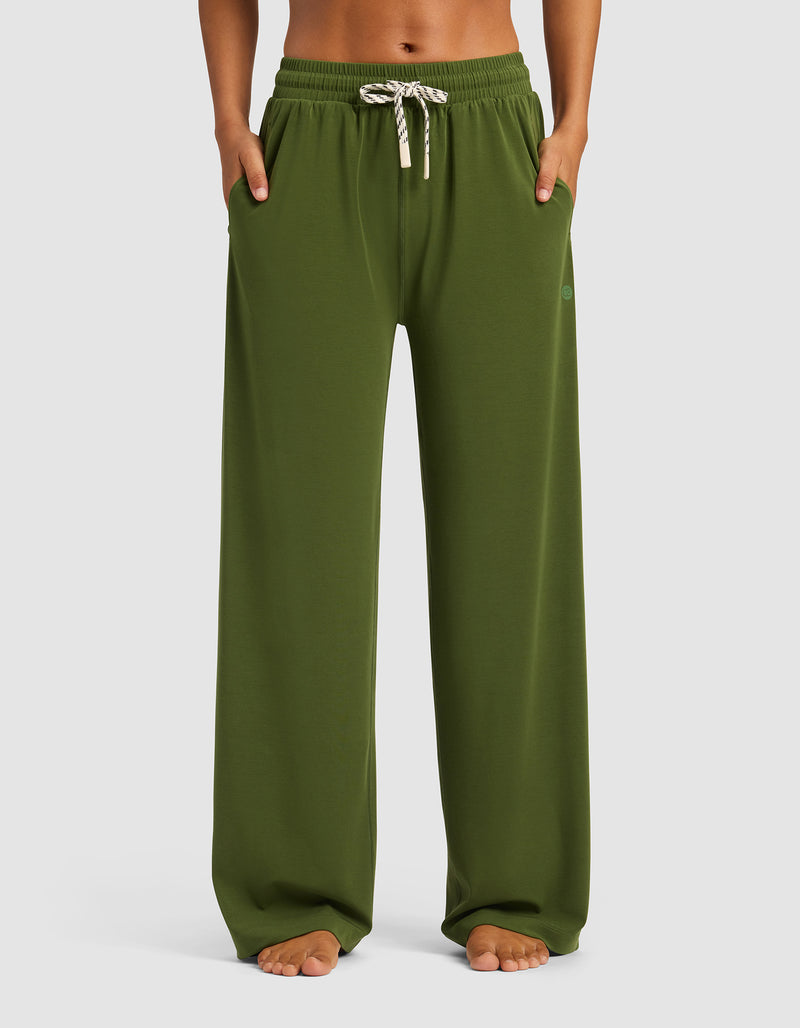 A woman stands barefoot with hands in pockets, wearing Cozy Earth's Women's Studio Wide Leg Pant in olive green, featuring an elastic waistband and white drawstring, against a simple background.