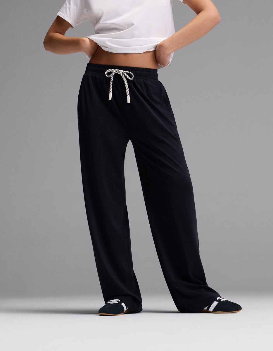 A person stands against a plain gray background wearing Cozy Earth’s Women’s Studio Wide Leg Pant in Jet Black, paired with a cropped white T-shirt and black slippers with white details. Their head and most of their arms are out of frame.