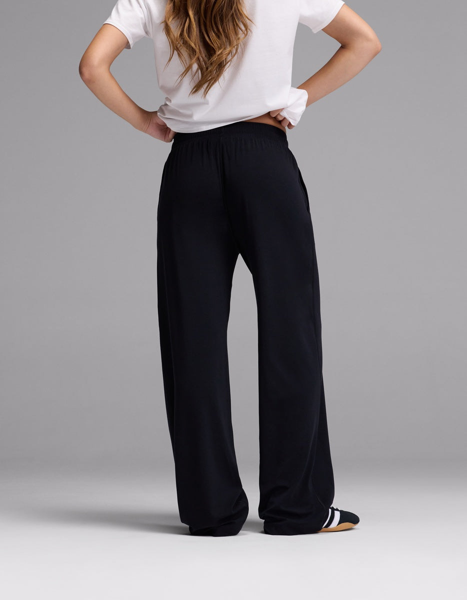 A person stands with hands on hips, facing away, wearing a white short-sleeve shirt and Cozy Earth Women’s Studio Wide Leg Pant in black. The background is plain gray.