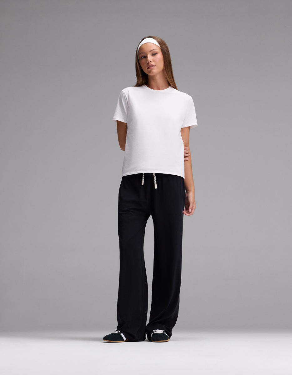 A woman stands against a gray background, wearing Cozy Earth’s Women’s Studio Wide Leg Pant, a plain white t-shirt, black slide sandals, and a white headband, with one arm behind her back.