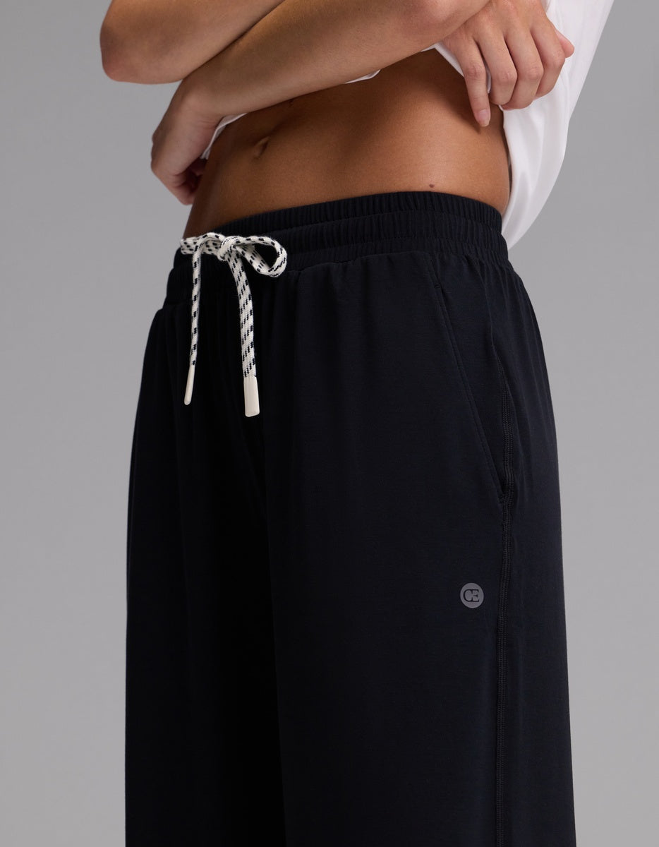 A woman is wearing Cozy Earth’s Women’s Studio Wide Leg Pant in black with a checkered drawstring at the waist and a white shirt, standing against a plain gray background.