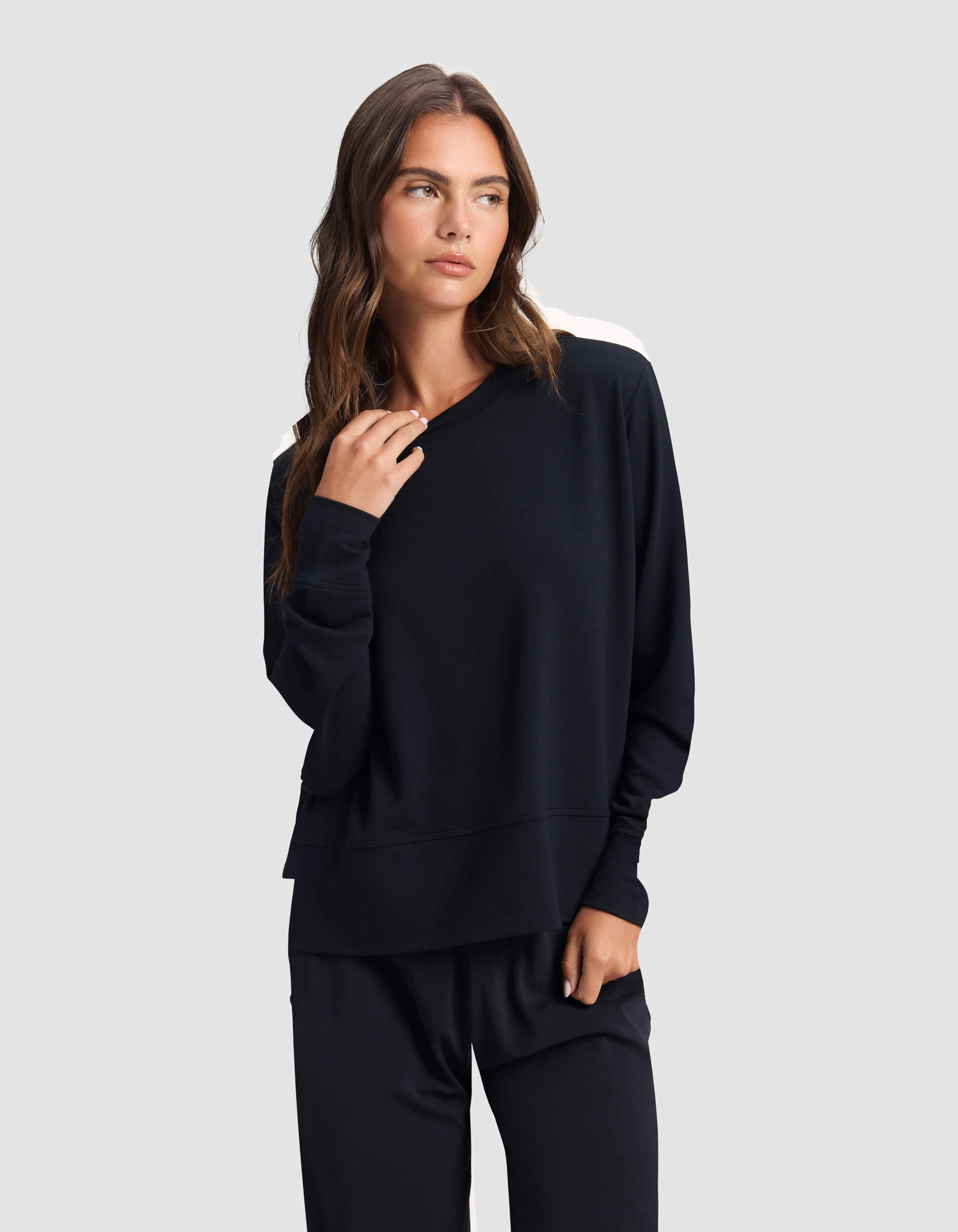 A woman with long brown hair wears the Cozy Earth Women's StudioLite Crewneck, a loose-fitting black sweatshirt with white shoulder accents, and matching black pants, standing against a plain light gray background.|Color:Jet Black