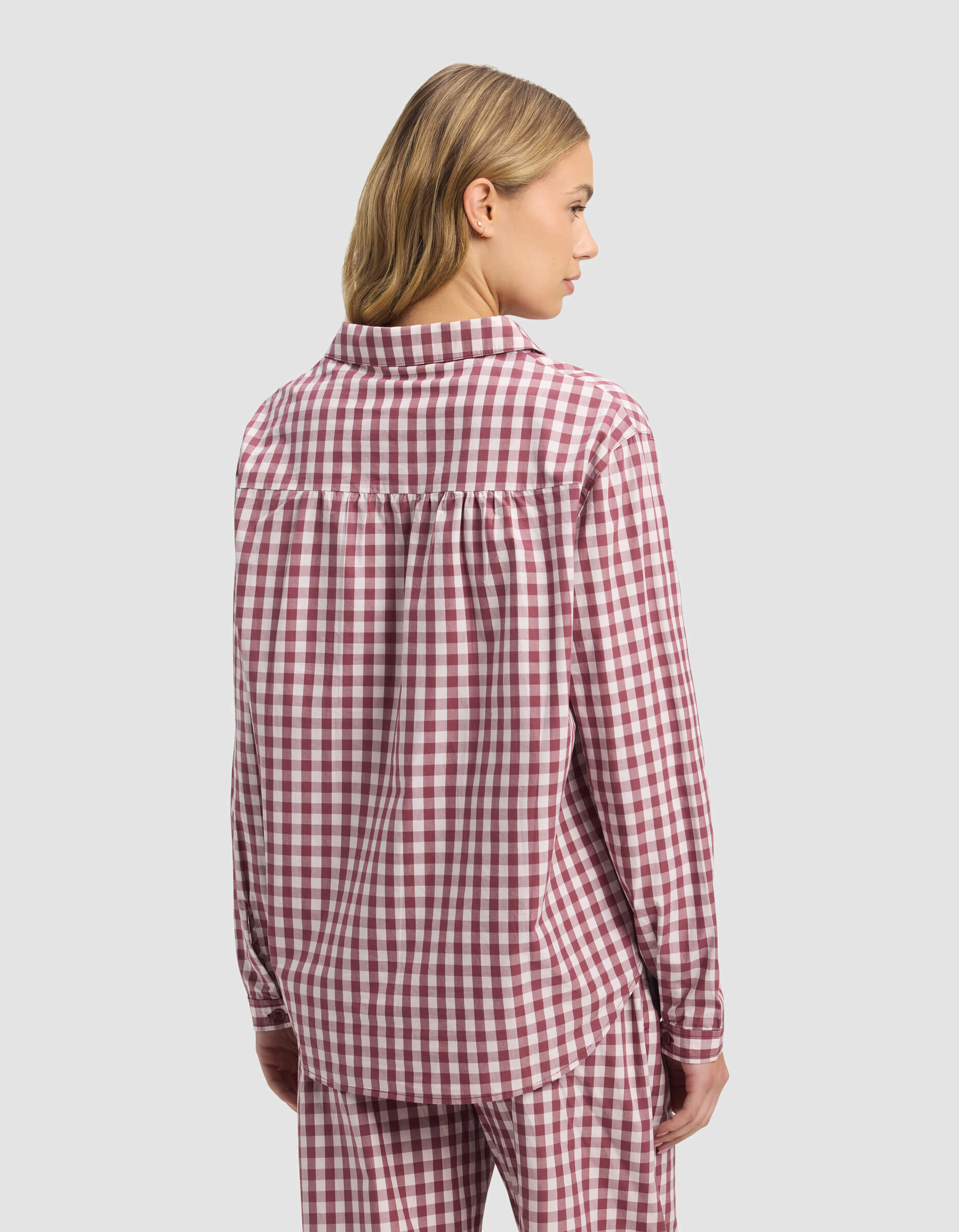 A woman with blonde hair wears the Cozy Earth Women's Sutton Long Sleeve Pajama Top in a red and white check, with matching pants, standing facing away against a plain light gray background.|Color:Mulberry Gingham