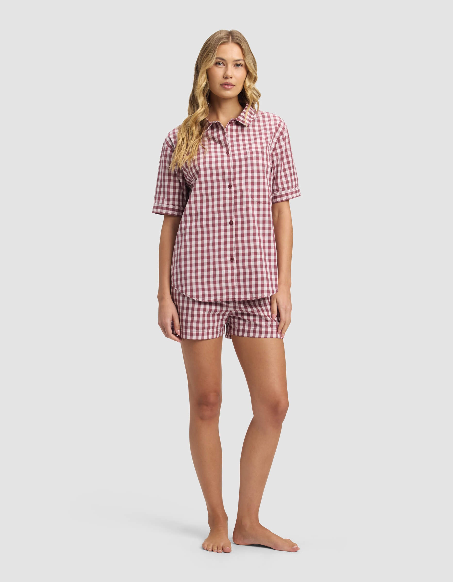 A woman stands barefoot against a plain light background wearing the Cozy Earth Women's Sutton Short Sleeve Pajama Set, featuring a red and white checkered pattern with a button-up shirt and matching shorts.|Color:Mulberry Gingham
