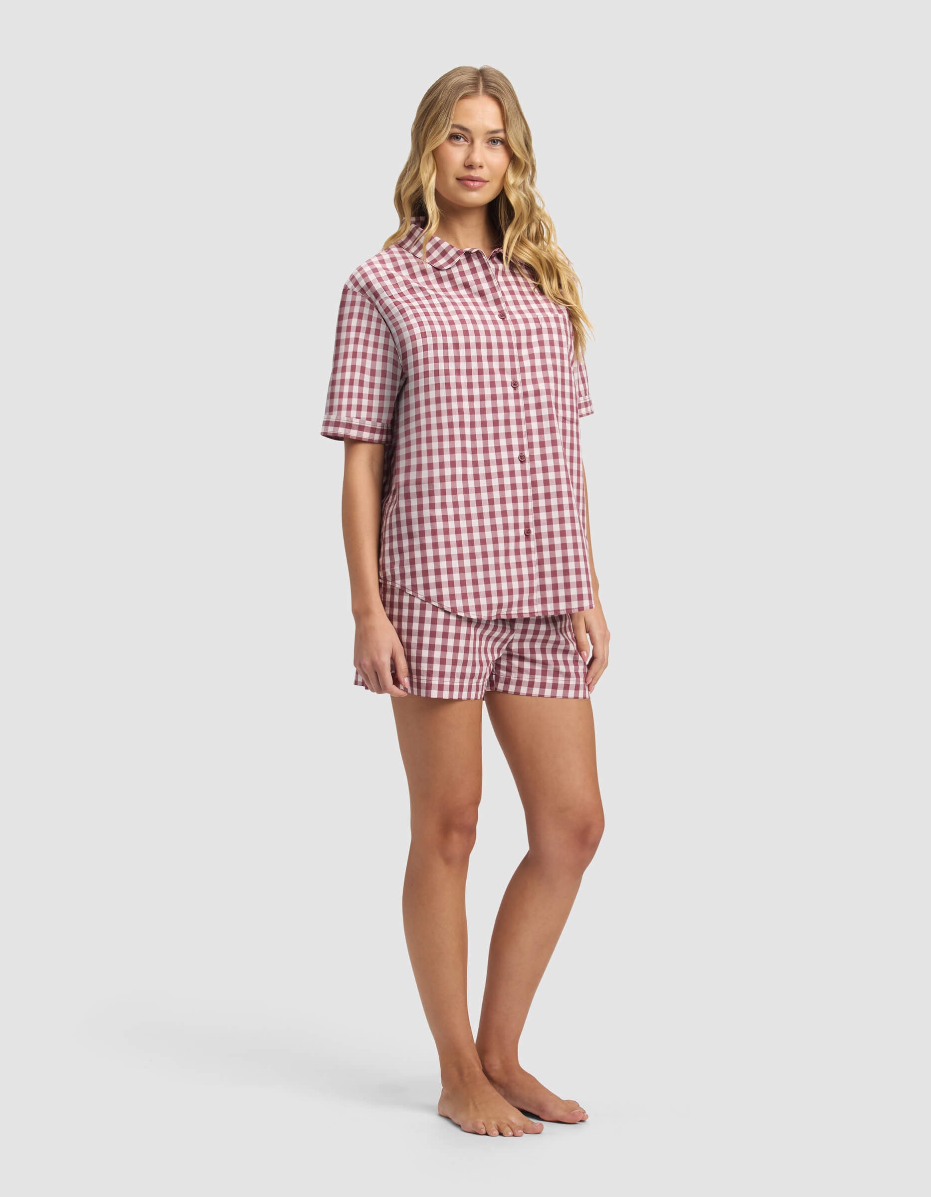 A woman stands barefoot against a light gray background, wearing Cozy Earth's Women's Sutton Short Sleeve Pajama Set in a red and white gingham pattern.|Color:Mulberry Gingham