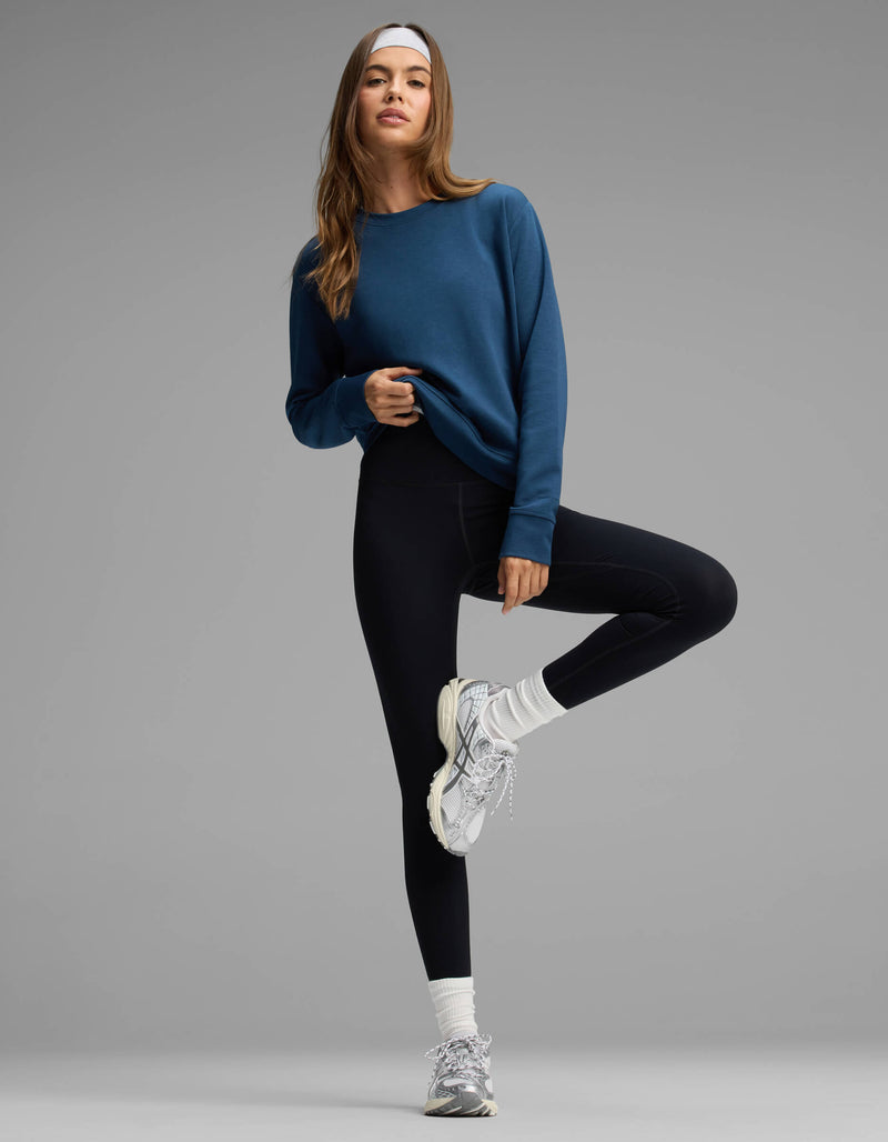 A woman stands on one leg against a neutral gray background, wearing the Cozy Earth Women's Versa Crewneck in blue, paired with black leggings, white sneakers, and a white headband.