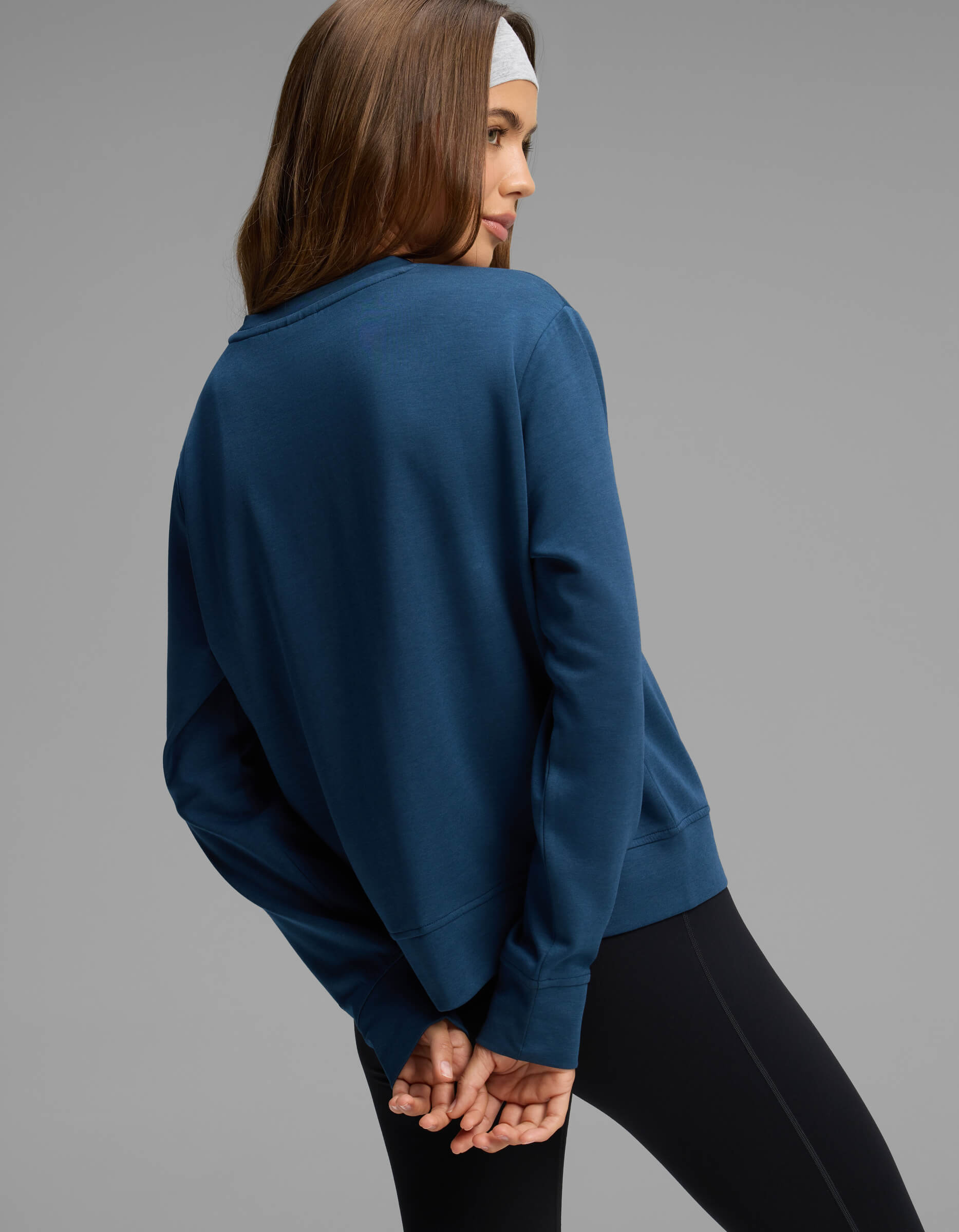A woman stands with her back turned and hands clasped behind her against a plain gray background, wearing the Cozy Earth Women's Versa Crewneck in blue.|Color:Obsidian