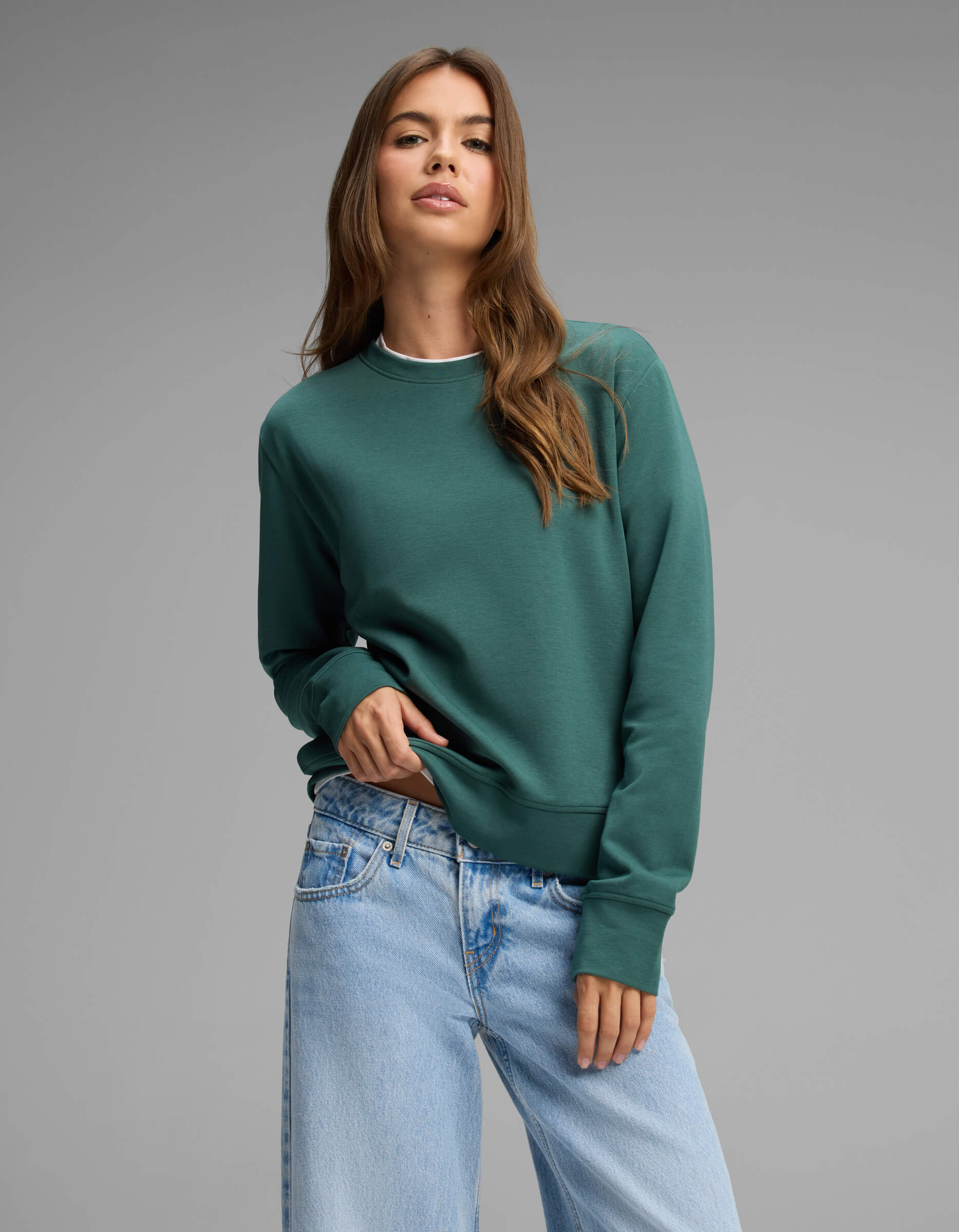 A woman with long brown hair wears the Cozy Earth Women's Versa Crewneck in green and light blue jeans, posing with one hand in her pocket against a plain gray background.