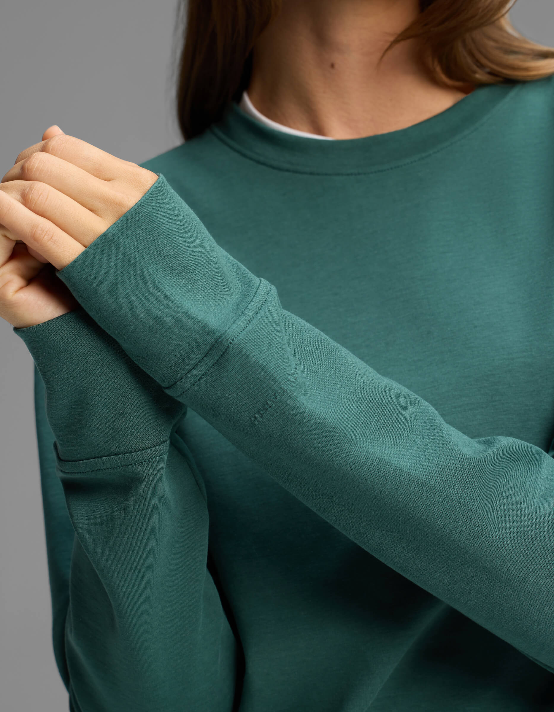 A person wearing the Cozy Earth Women's Versa Crewneck in teal clasps their hands together in front of their chest. The face is not visible, and the background is plain gray.