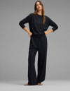 A woman stands against a gray background wearing the Cozy Earth Women's Versa Crewneck & Straight Leg Pant Set in black, paired with sandals. She has long brown hair and poses confidently with her hands on her hips, looking at the camera.|Color:Jet Black