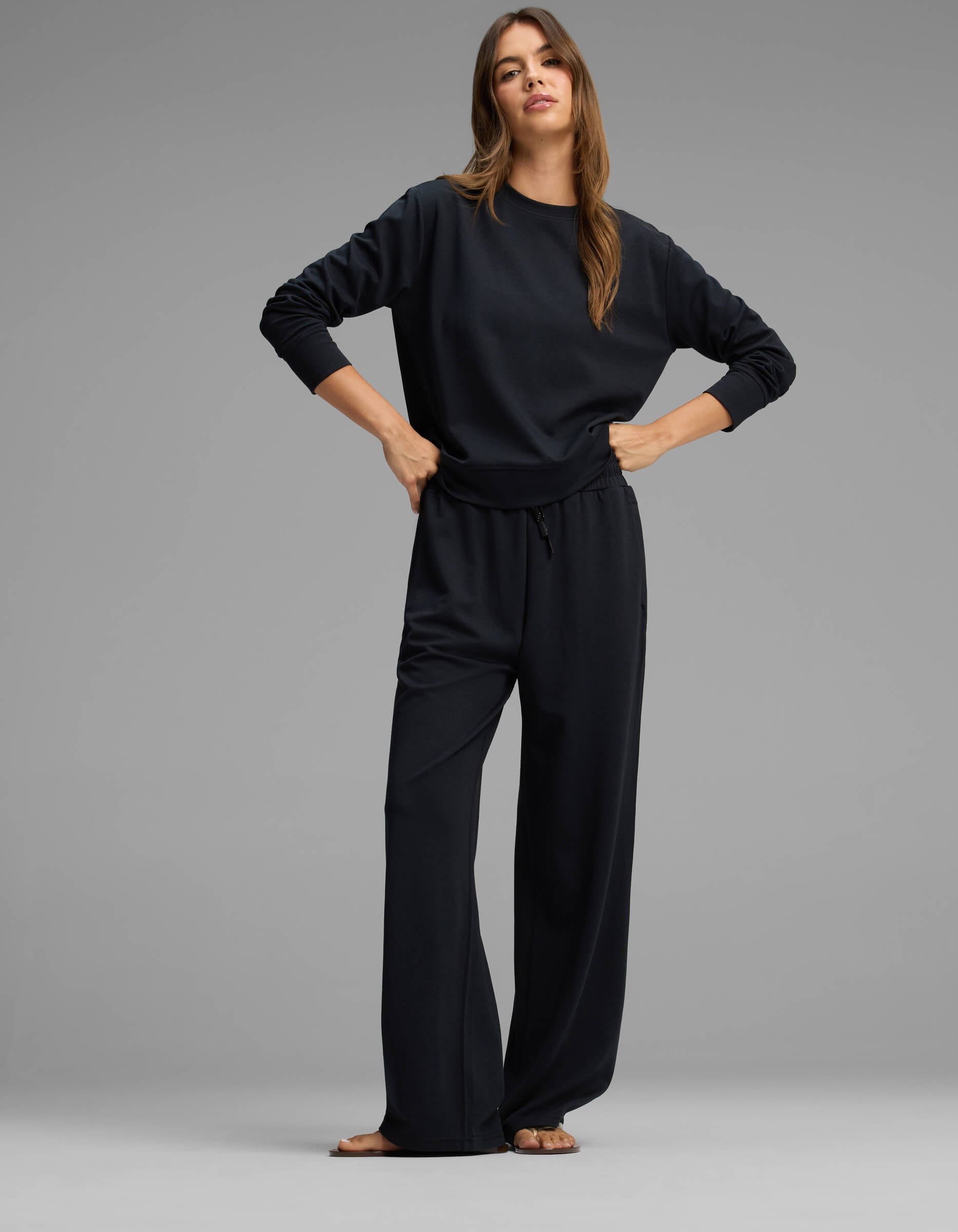 A woman stands against a gray background wearing the Cozy Earth Women's Versa Crewneck & Straight Leg Pant Set in black, paired with sandals. She has long brown hair and poses confidently with her hands on her hips, looking at the camera.|Color:Jet Black