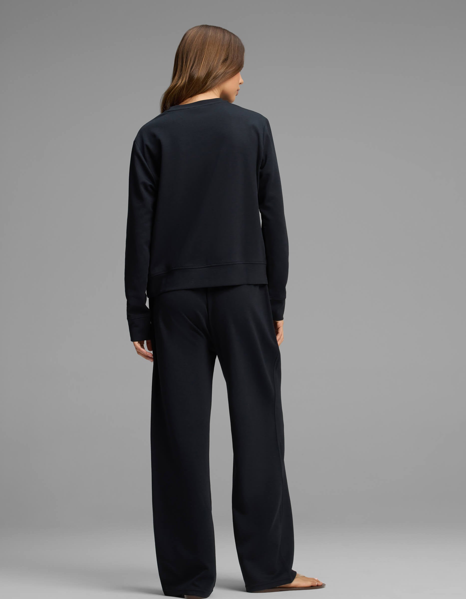 A woman with long brown hair stands barefoot, facing away from the camera, wearing the Cozy Earth Women's Versa Crewneck & Straight Leg Pant Set in black against a plain gray background.