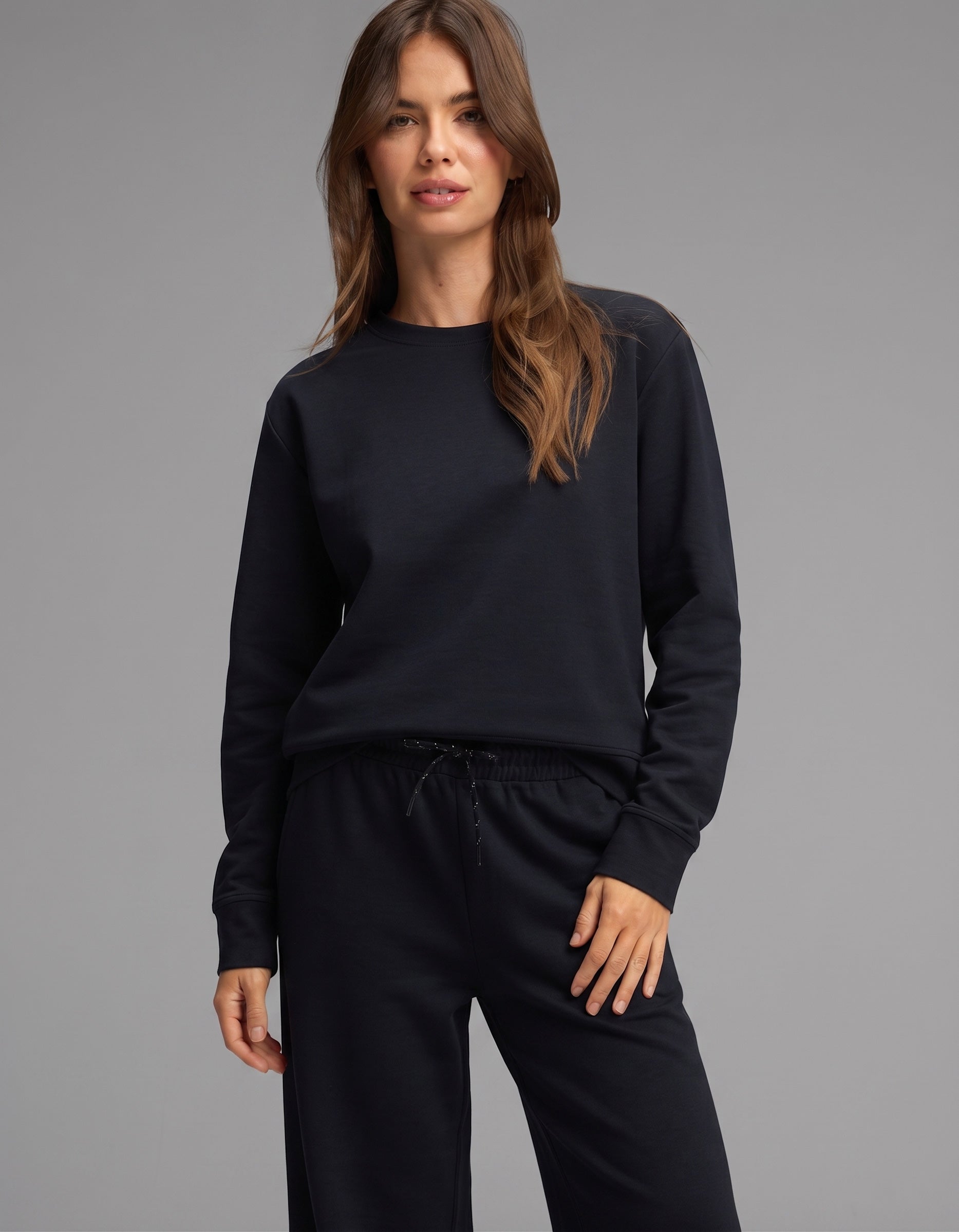A woman with long brown hair stands against a plain gray background, looking at the camera while wearing the Cozy Earth Women's Versa Crewneck & Wide Leg Pant Set in black.
