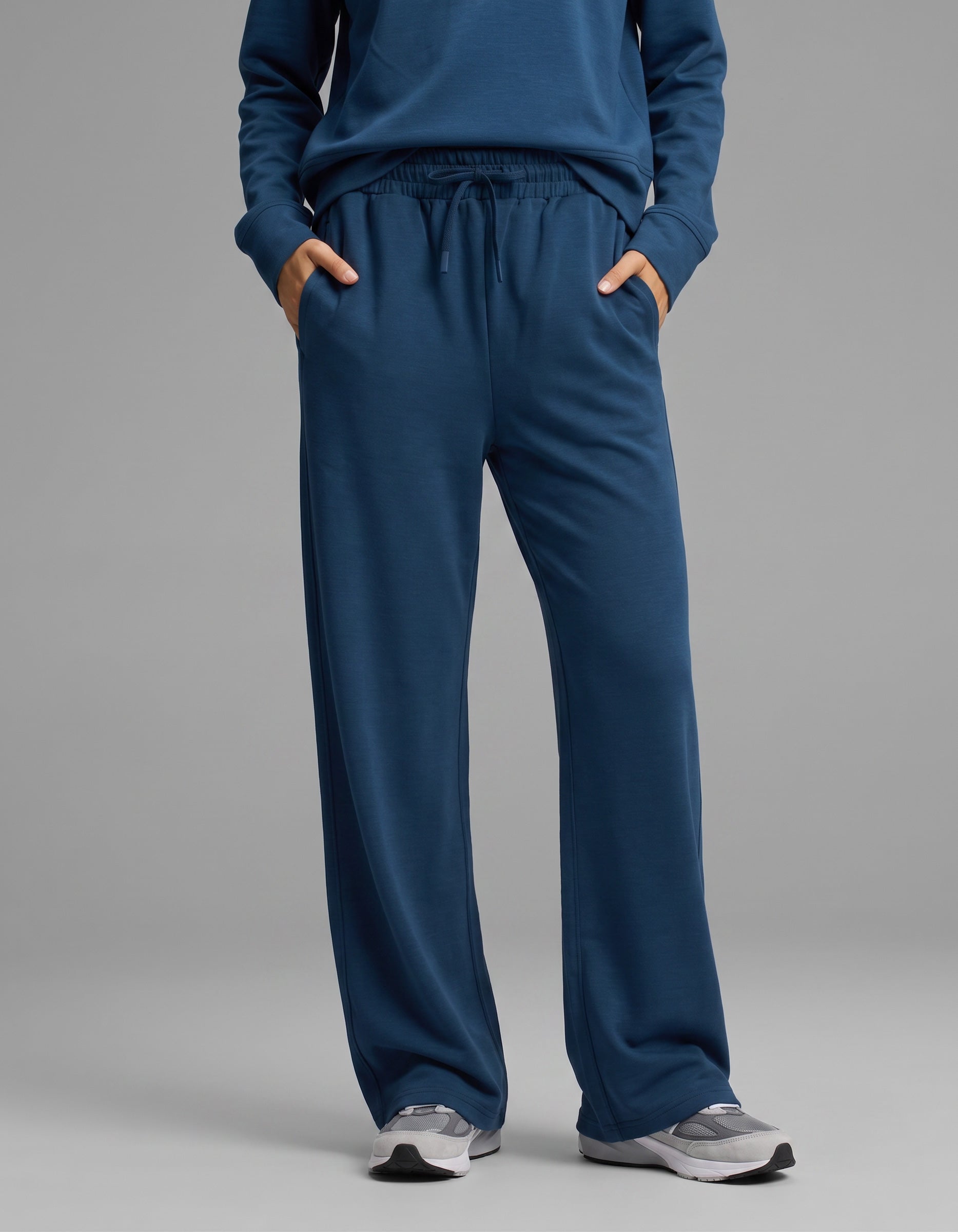 A person is shown from the waist down wearing the Cozy Earth Women's Versa Crewneck & Wide Leg Pant Set in blue, with hands in the pockets of the wide leg pants. They are also wearing gray athletic shoes, standing against a plain light gray background.