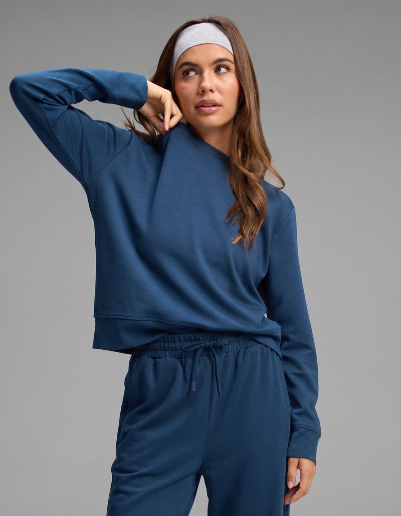 A woman stands against a plain background, wearing the Cozy Earth Women's Versa Crewneck & Wide Leg Pant Set in blue, paired with a light gray headband. She raises one arm to touch her face.