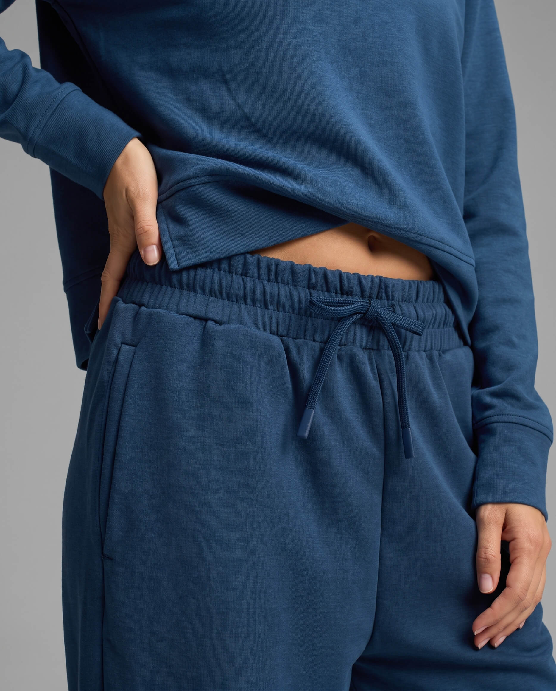 A person models the Cozy Earth Women's Versa Crewneck & Wide Leg Pant Set in blue, with one hand tucked into a pocket and the other lifting the sweatshirt slightly to reveal part of their torso.