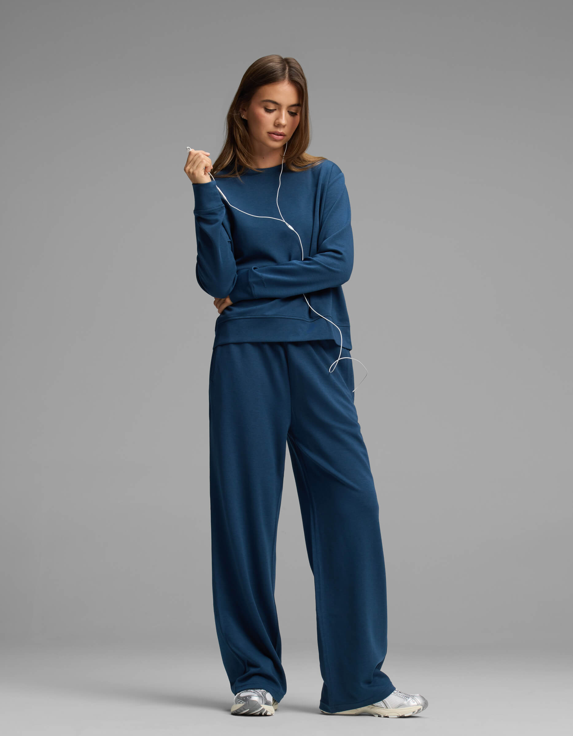 A woman wears the Cozy Earth Women's Versa Crewneck & Straight Leg Pant Set with sneakers, standing against a gray background and holding wired earphones with a relaxed expression.