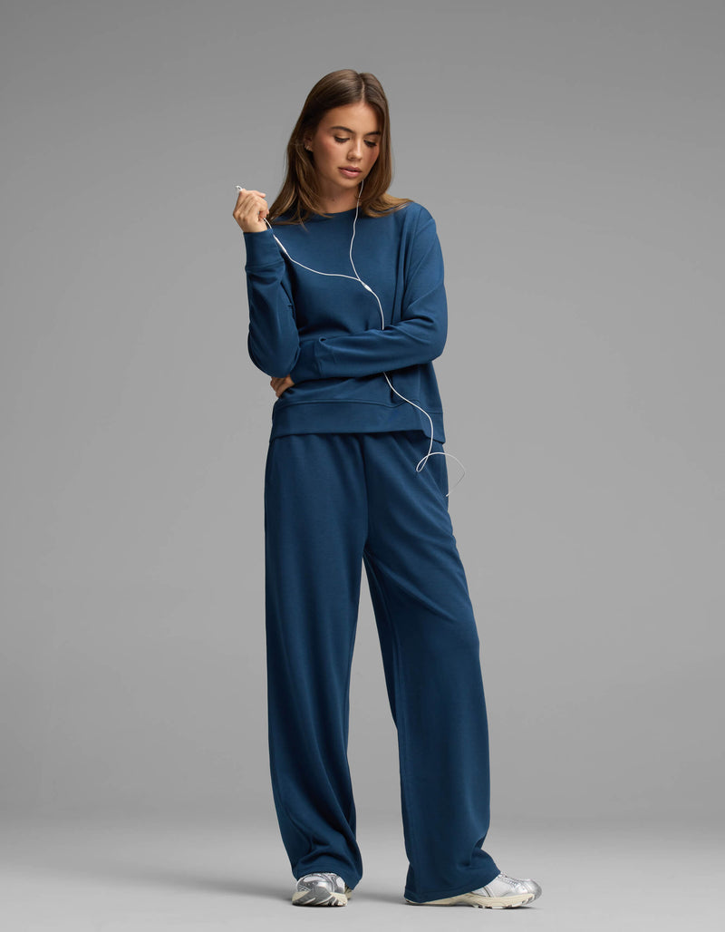 A woman wears the Cozy Earth Women's Versa Crewneck & Straight Leg Pant Set with sneakers, standing against a gray background and holding wired earphones with a relaxed expression.
