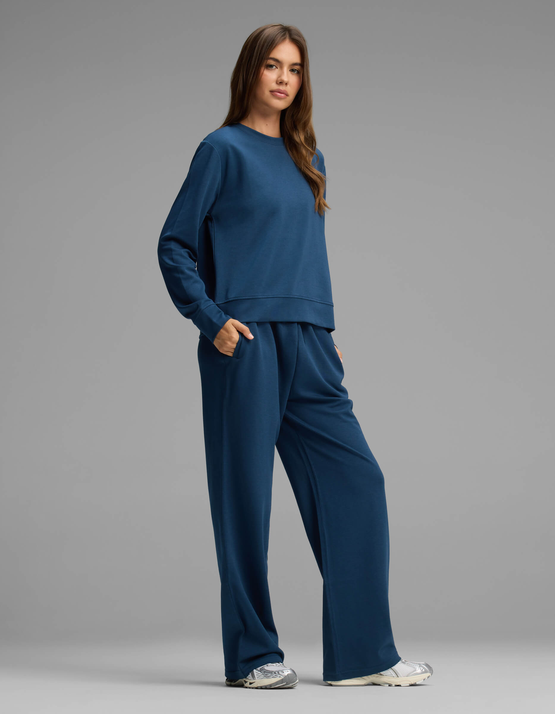 A person stands against a plain gray background, wearing the Cozy Earth Women's Versa Crewneck & Straight Leg Pant Set in matching blue with hands in pockets, paired with white sneakers.|Color:Obsidian