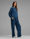 A person stands against a plain gray background, wearing the Cozy Earth Women's Versa Crewneck & Straight Leg Pant Set in matching blue with hands in pockets, paired with white sneakers.|Color:Obsidian