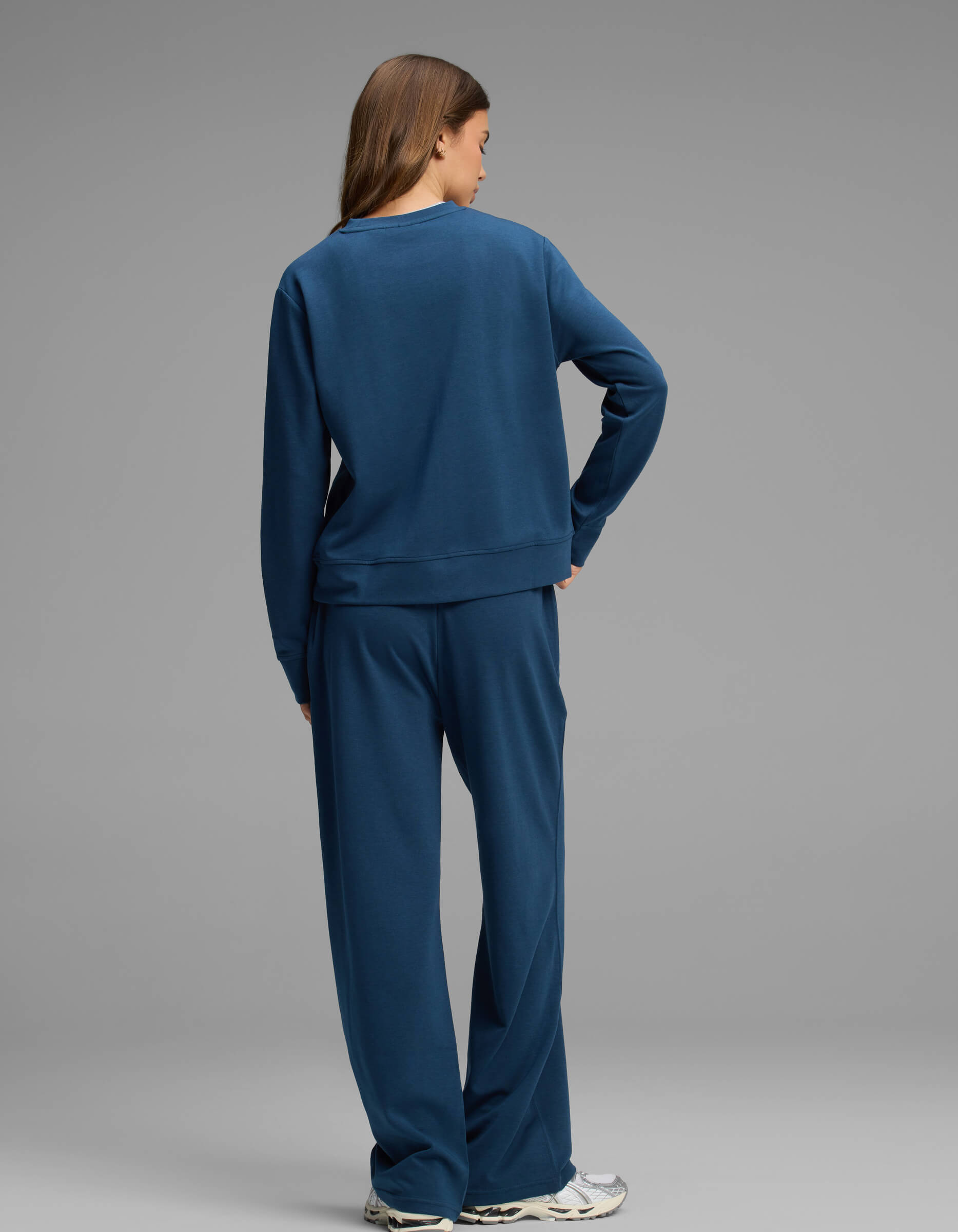 A person stands facing away, wearing the Cozy Earth Women's Versa Crewneck & Straight Leg Pant Set in blue with white sneakers, against a plain gray background.|Color:Obsidian