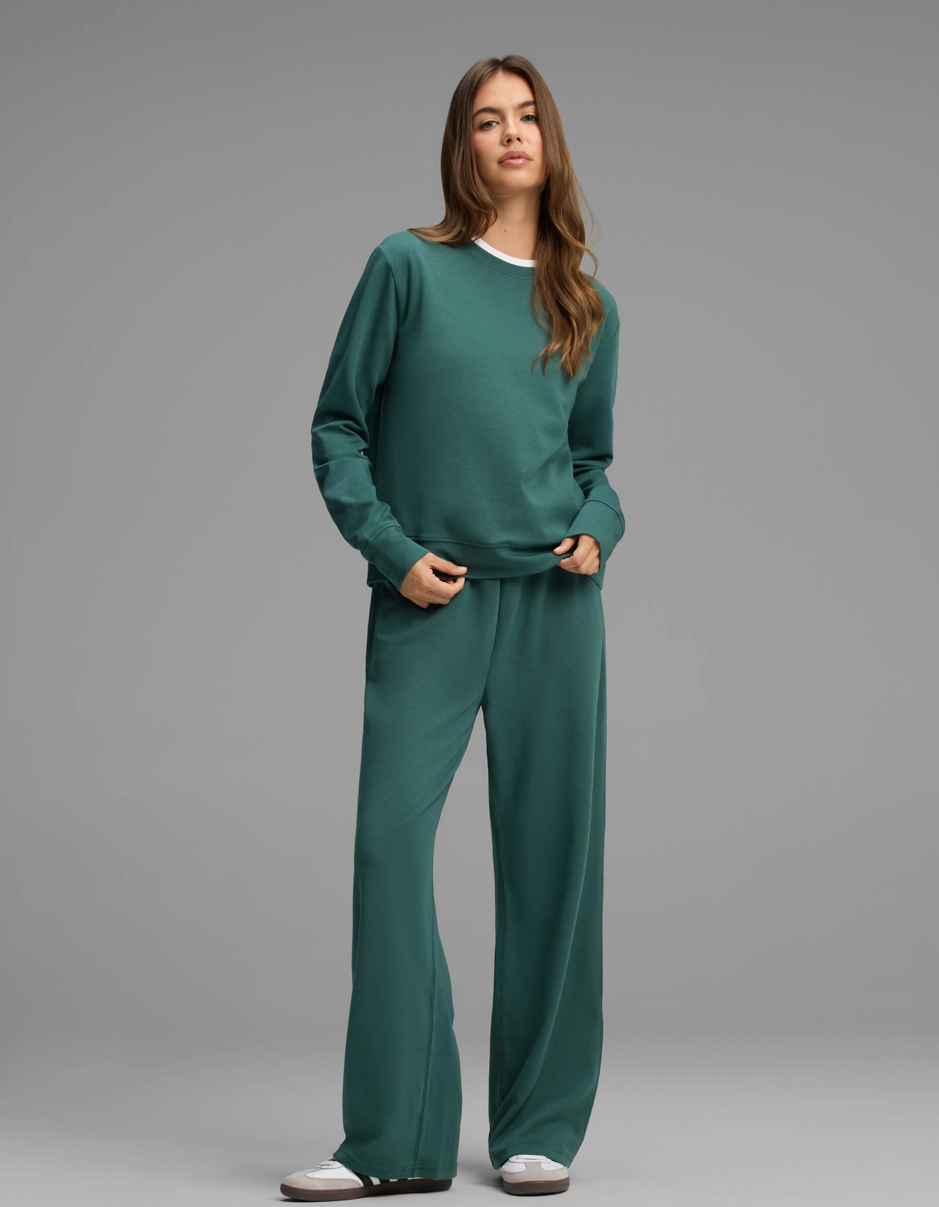 A person stands against a gray background, wearing the Cozy Earth Women's Versa Crewneck & Straight Leg Pant Set in teal, paired with white sneakers.