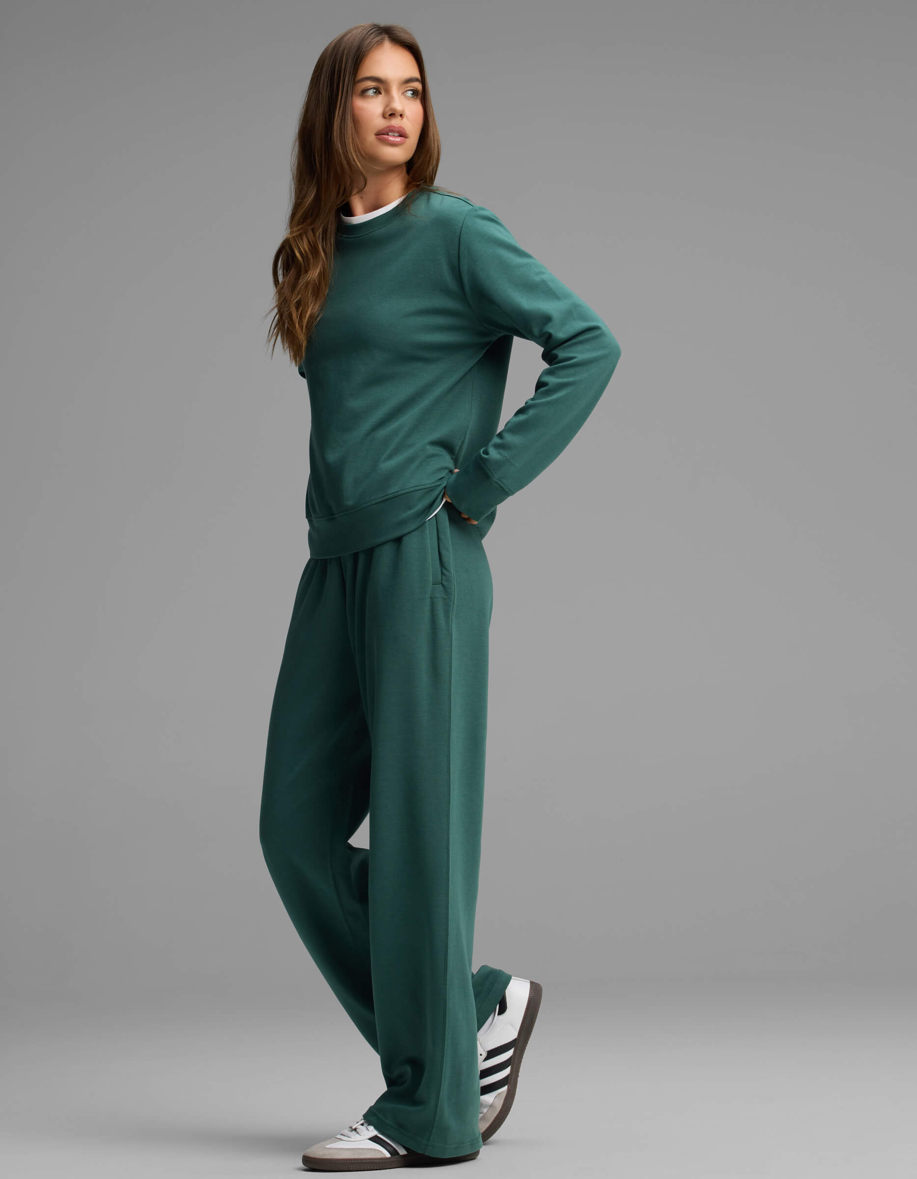 A person stands sideways against a plain gray background, modeling the Cozy Earth Women's Versa Crewneck & Straight Leg Pant Set in dark green with sneakers.
