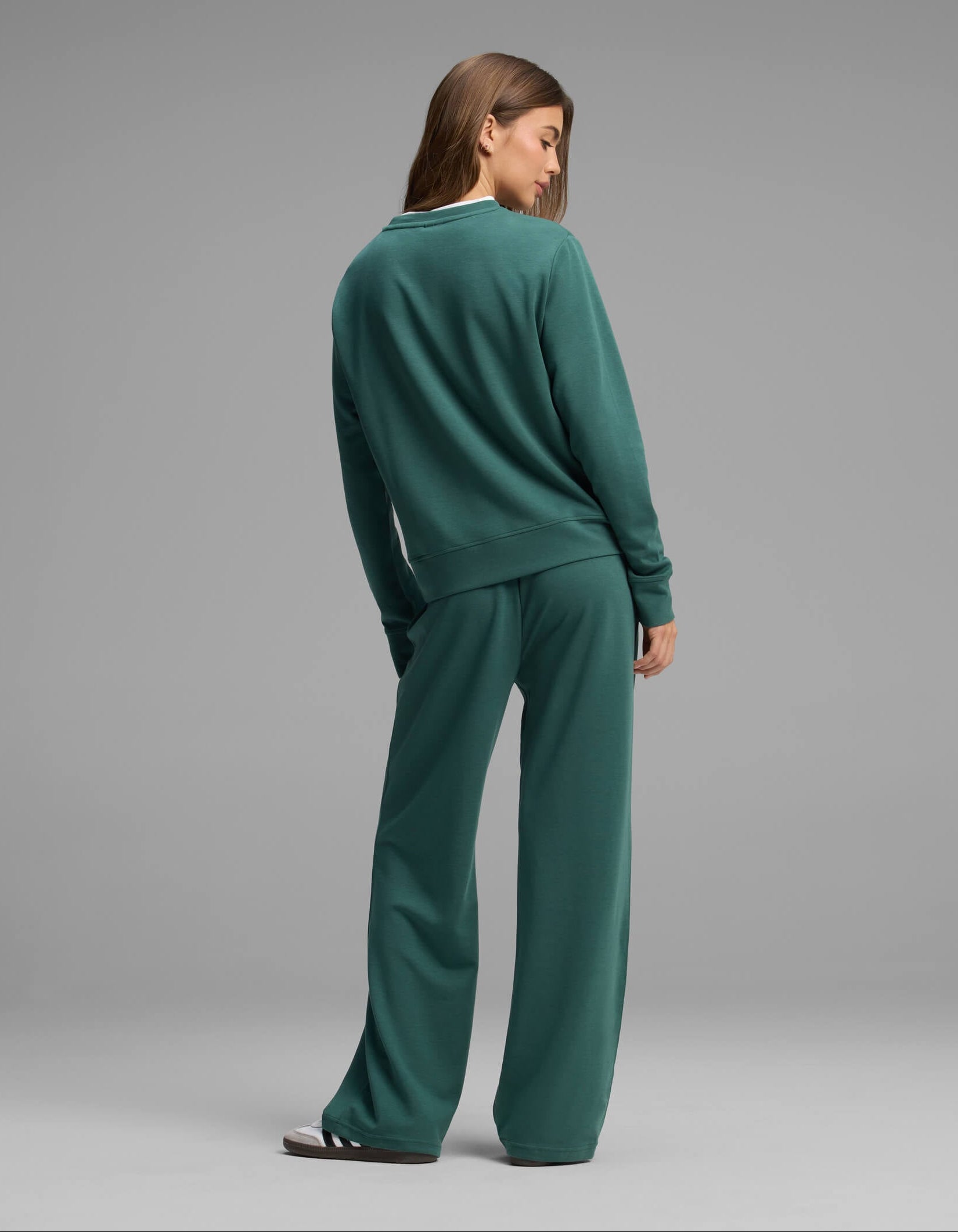 A woman stands facing away, wearing the Cozy Earth Women's Versa Crewneck & Straight Leg Pant Set in green. She looks over her shoulder against a plain gray background.