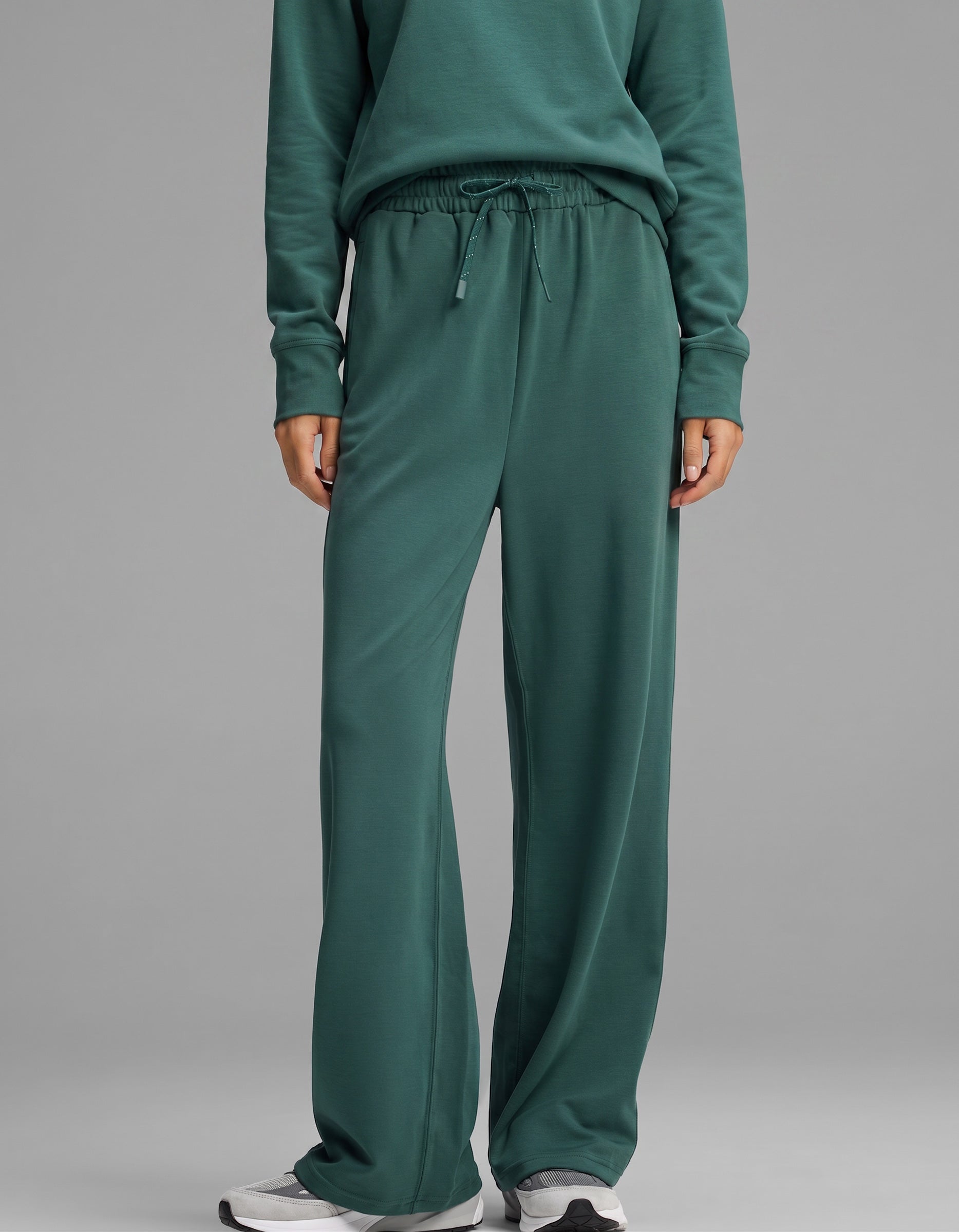 A person is shown from the waist down wearing the Cozy Earth Women's Versa Crewneck & Wide Leg Pant Set in dark green, featuring loose-fitting wide leg pants with a drawstring waist and a matching sweatshirt, paired with white sneakers.