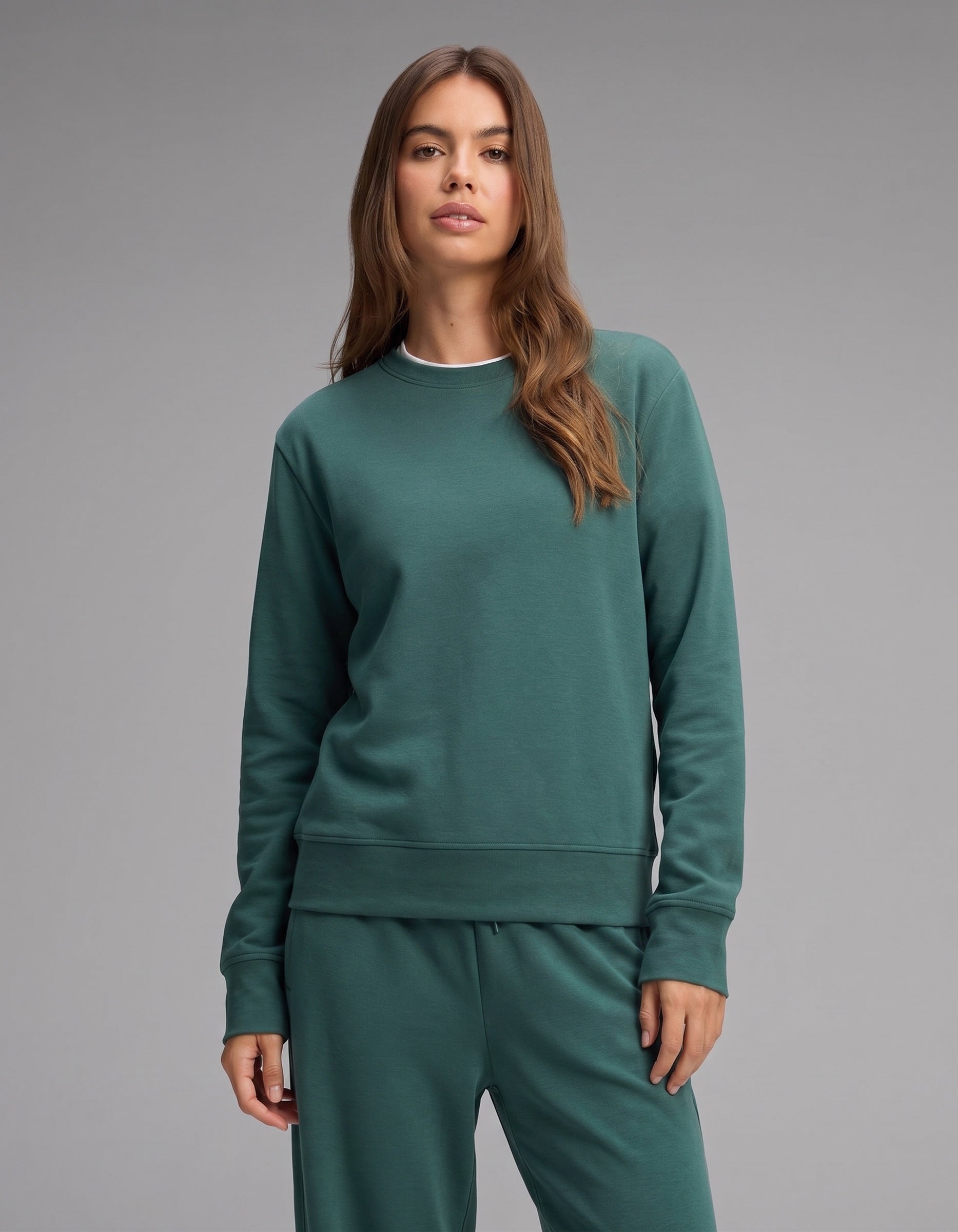 A woman with long brown hair stands against a plain gray background, wearing the Women's Versa Crewneck & Wide Leg Pant Set by Cozy Earth in green.