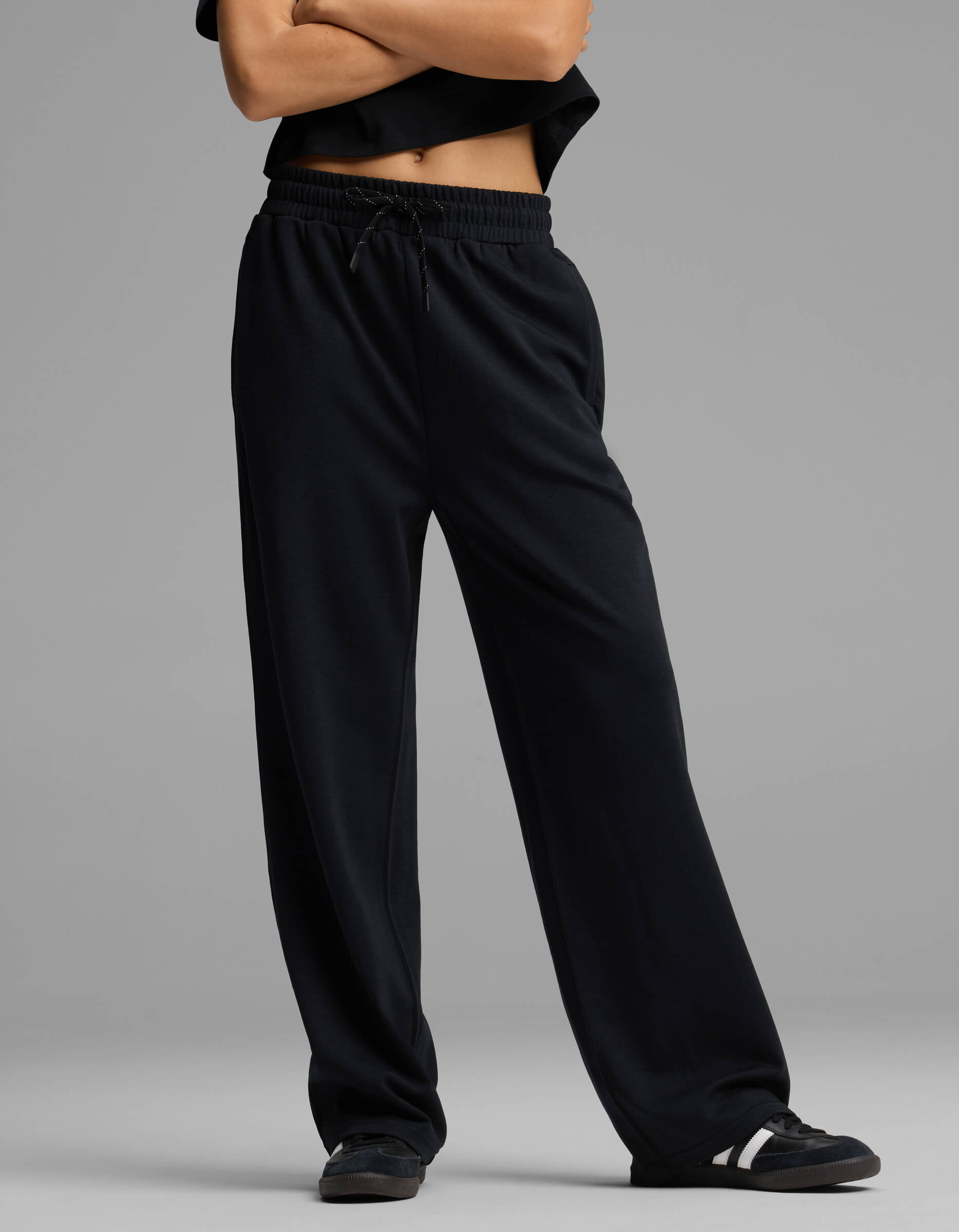 A person stands with arms crossed, dressed in Cozy Earth's Women's Versa Straight Leg Pant, paired with a black top and black sneakers with white accents against a plain gray background. Their face is not visible in the image.|Color:Jet Black