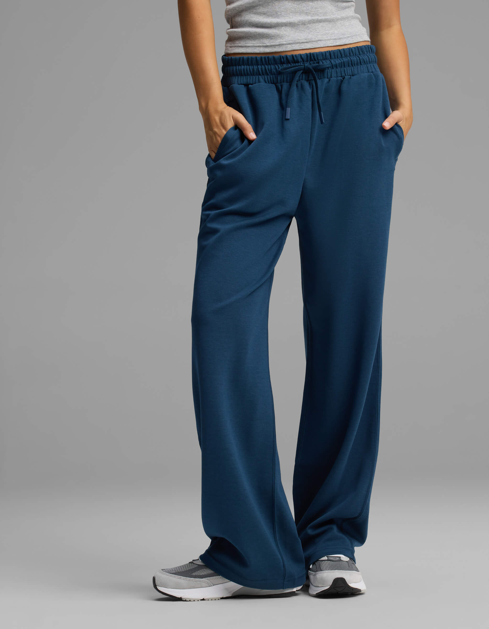 A person is wearing the Cozy Earth Women's Versa Straight Leg Pant in blue, paired with a gray tank top and gray sneakers, standing with hands in their pockets against a plain gray background.|Color:Obsidian