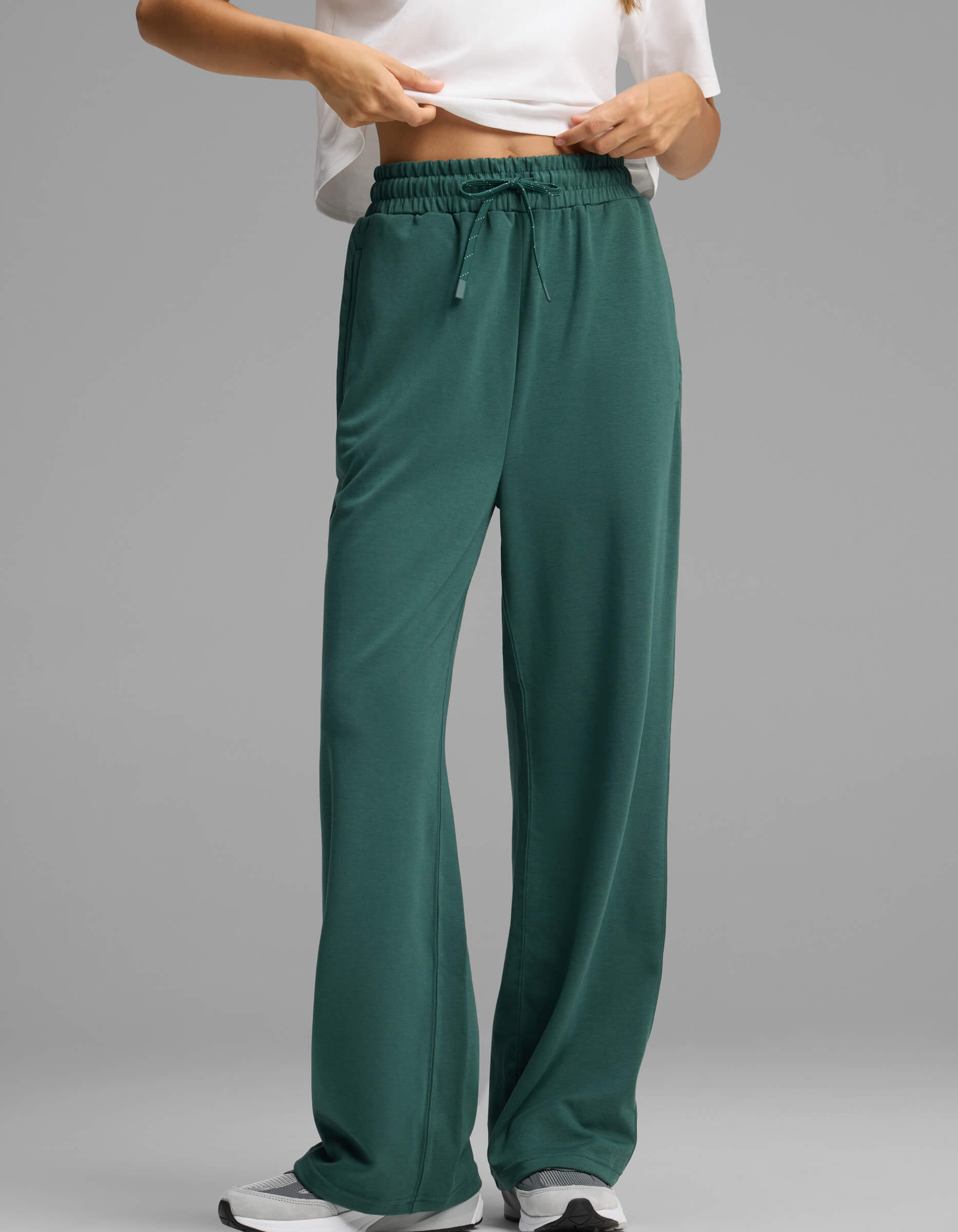 A model showcases the lower half of the Women's Versa Straight Leg Pant by Cozy Earth in green, paired with a white shirt and white sneakers, standing against a plain grey background and seen adjusting the waistband.|Color:Uniform