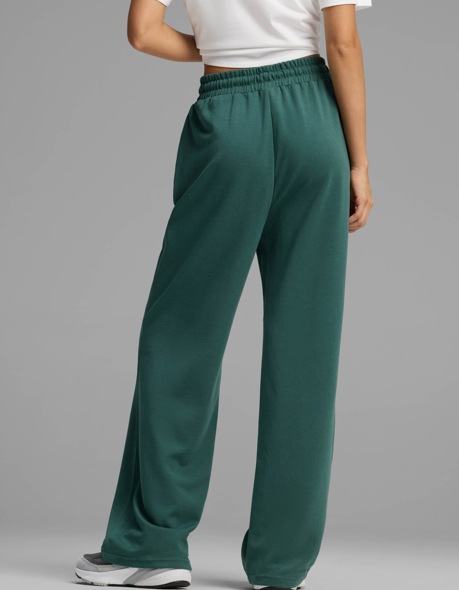 A person is shown from behind standing on a gray background, wearing Cozy Earth Women's Versa Straight Leg Pant in green, paired with a white short-sleeve shirt and white sneakers.