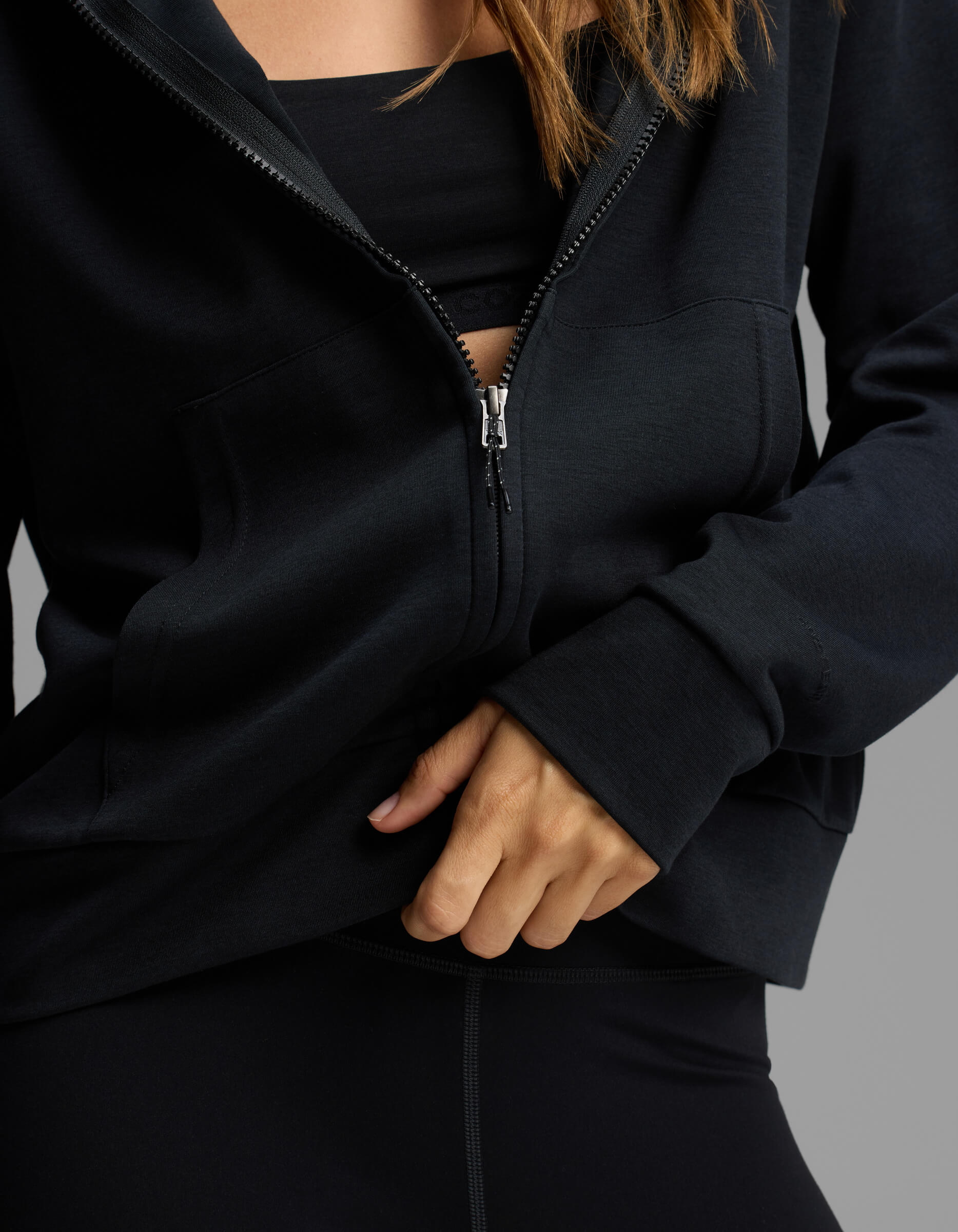 A person wearing the Cozy Earth Women's Versa Full-Zip Hoodie in black, partially unzipped to reveal a black sports bra and part of their hand and upper body, poses against a neutral background.