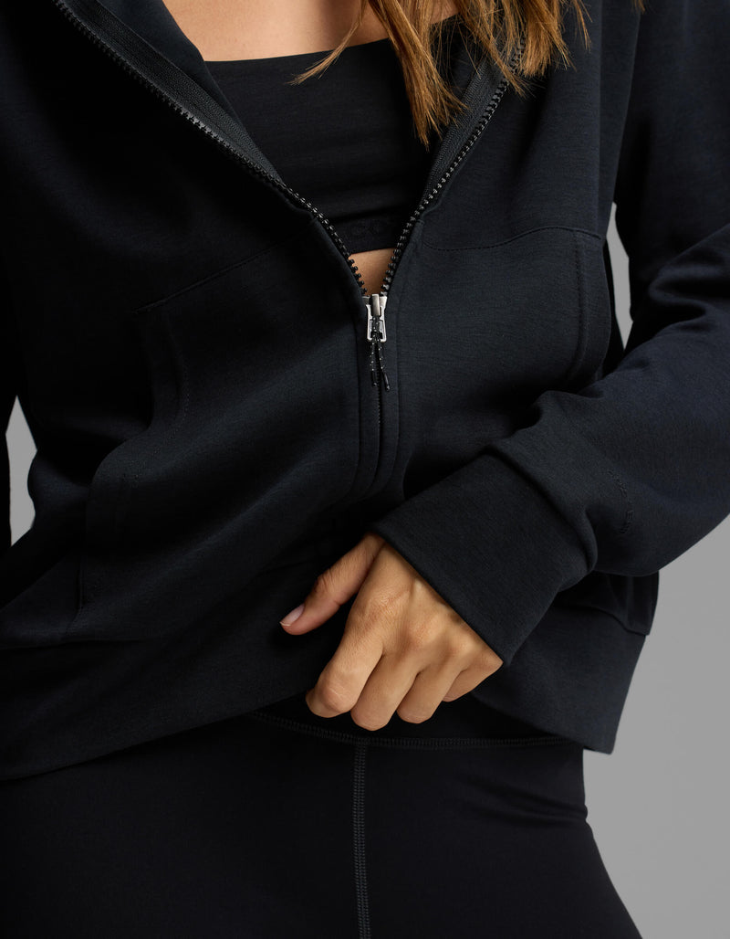 A person wearing the Cozy Earth Women's Versa Full-Zip Hoodie in black, partially unzipped to reveal a black sports bra and part of their hand and upper body, poses against a neutral background.