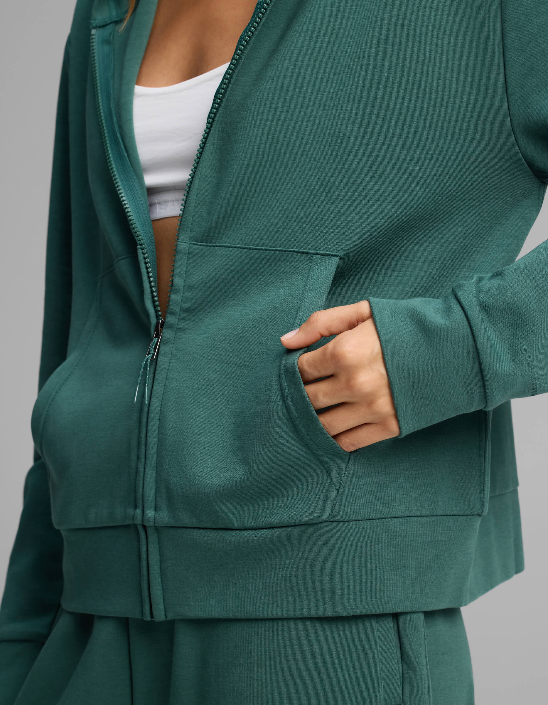 A person wearing the Cozy Earth Women's Versa Full-Zip Hoodie in green with their right hand in the front pocket; a white top is partially visible underneath.