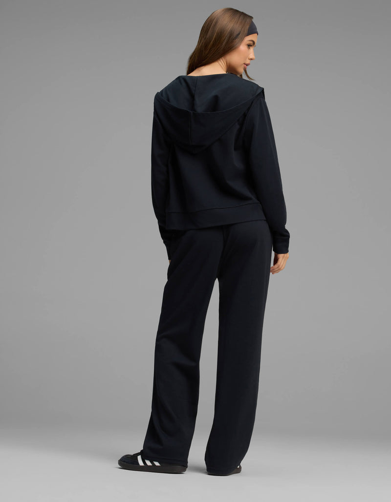 A woman stands facing away, wearing the Cozy Earth Women's Versa Zip-Down Hoodie & Straight Leg Pant Set in black, paired with black slide sandals featuring white stripes, against a plain grey background.