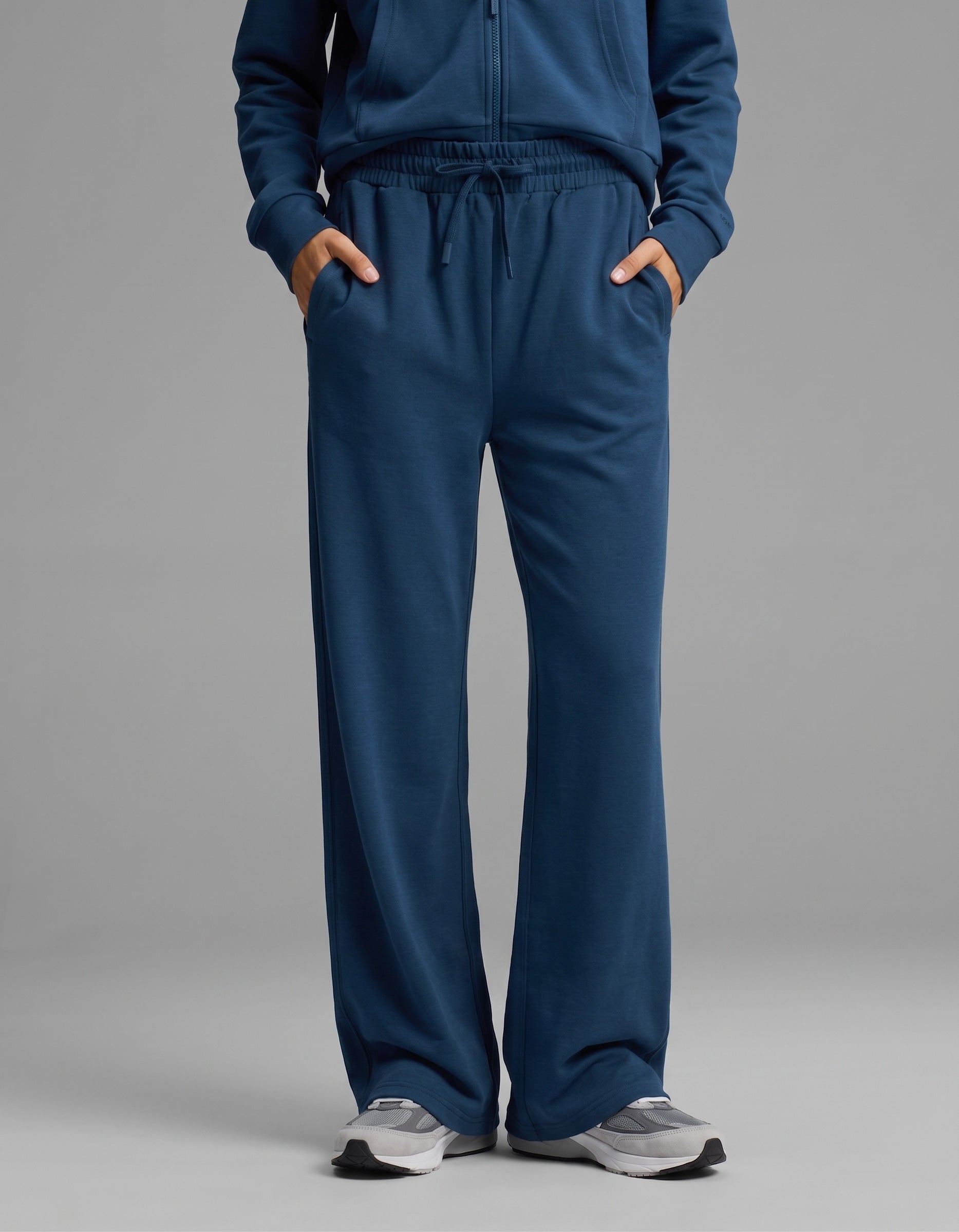 A person models Cozy Earth's Women's Versa Zip-Down Hoodie & Straight Leg Pant Set in blue, paired with gray sneakers, standing against a plain gray background with hands in pockets.