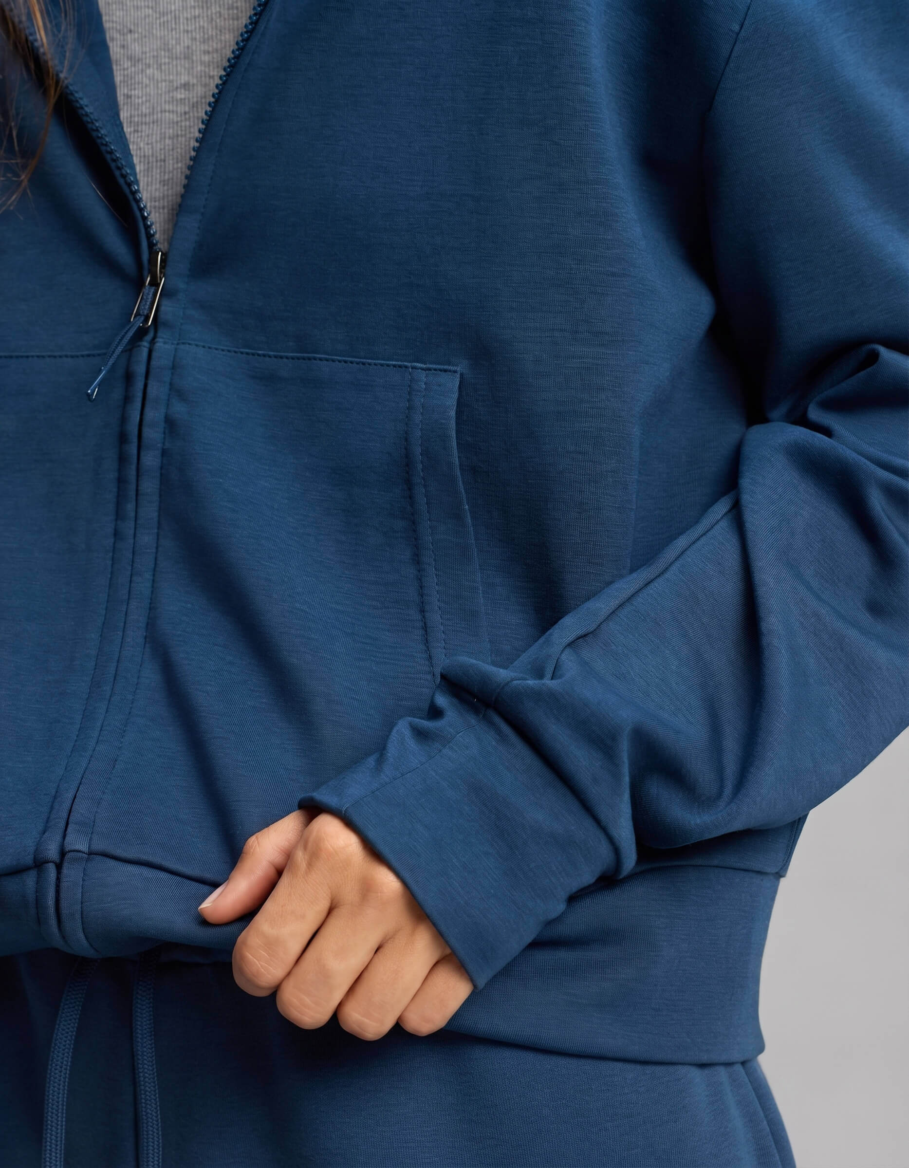 A person wearing the Cozy Earth Women's Versa Zip-Down Hoodie in blue, layered over a gray shirt, with one hand holding the hoodie near the pocket.