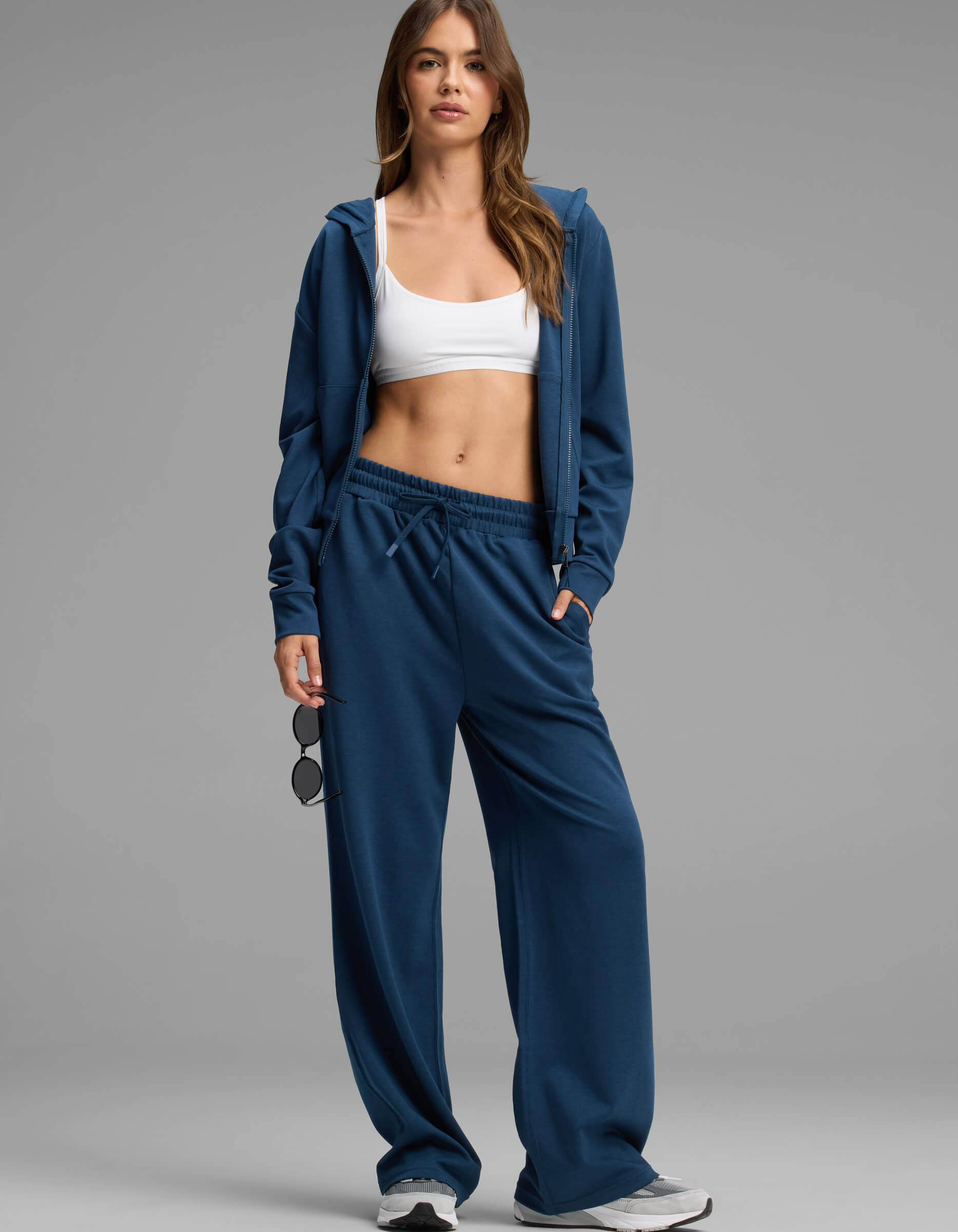 A woman stands wearing the Cozy Earth Women's Versa Zip-Down Hoodie & Straight Leg Pant Set in blue, paired with a white sports bra and sneakers. She holds sunglasses in one hand and gazes at the camera with a neutral expression against a plain background.
