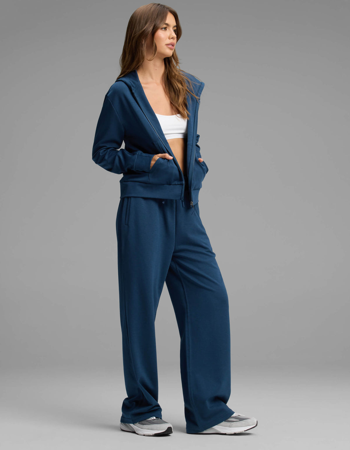 A woman stands against a plain gray background wearing the Cozy Earth Women's Versa Zip-Down Hoodie & Straight Leg Pant Set in blue, paired with a white crop top and white sneakers.|Color:Obsidian